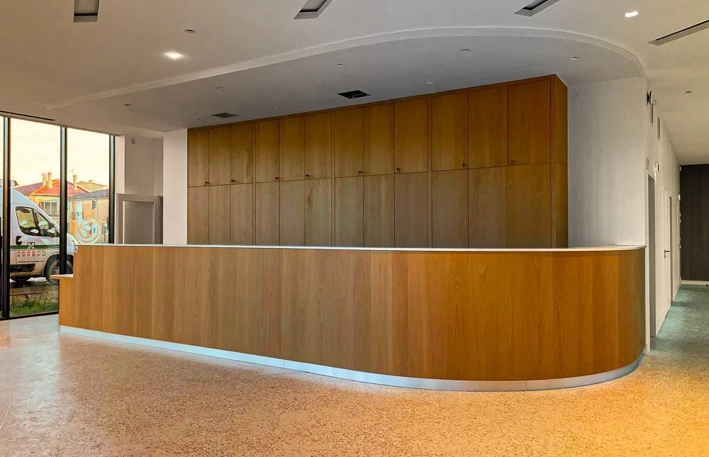 Un comptoir en bois dans un intérieur moderne, avec un mur en bois derrière, grandes fenêtres laissant entrer de la lumière naturelle.