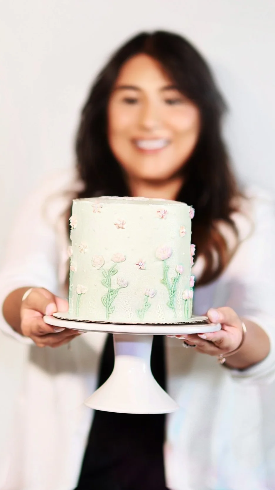 Woman with long dark hair smiling, holding a floral decorated cake on a white cake stand.