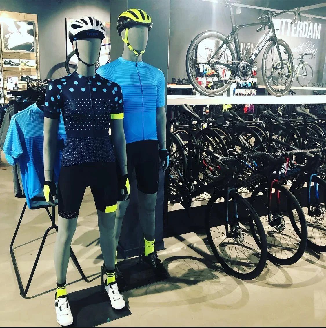 Display of athletic cycling mannequins in colorful jerseys and shorts, wearing helmets, surrounded by bicycles and cycling accessories in a sports store.