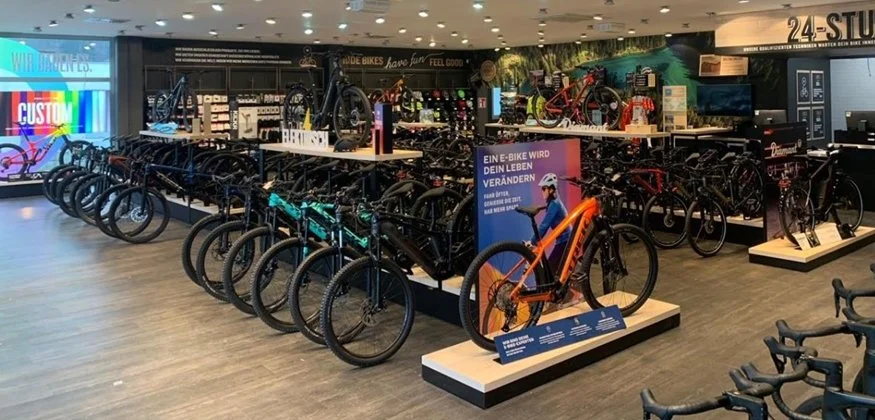 An indoor bike shop displays various bicycles, including mountain bikes, road bikes, and electric bikes, arranged on racks and platforms.