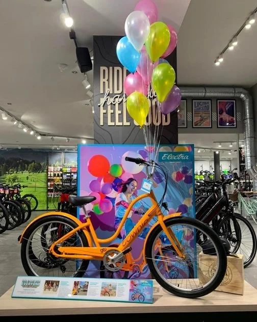 Orange electric bicycle displayed on a stand with colorful balloons attached to the handlebars inside a retail store.