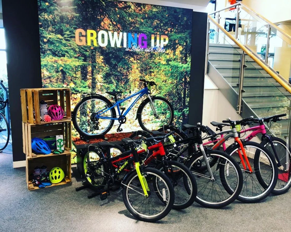 Indoor display of bikes with a woodland-themed sign that says 'Growing Up' and 'Better with Biki,' with helmets and supplies on wooden shelves to the side.