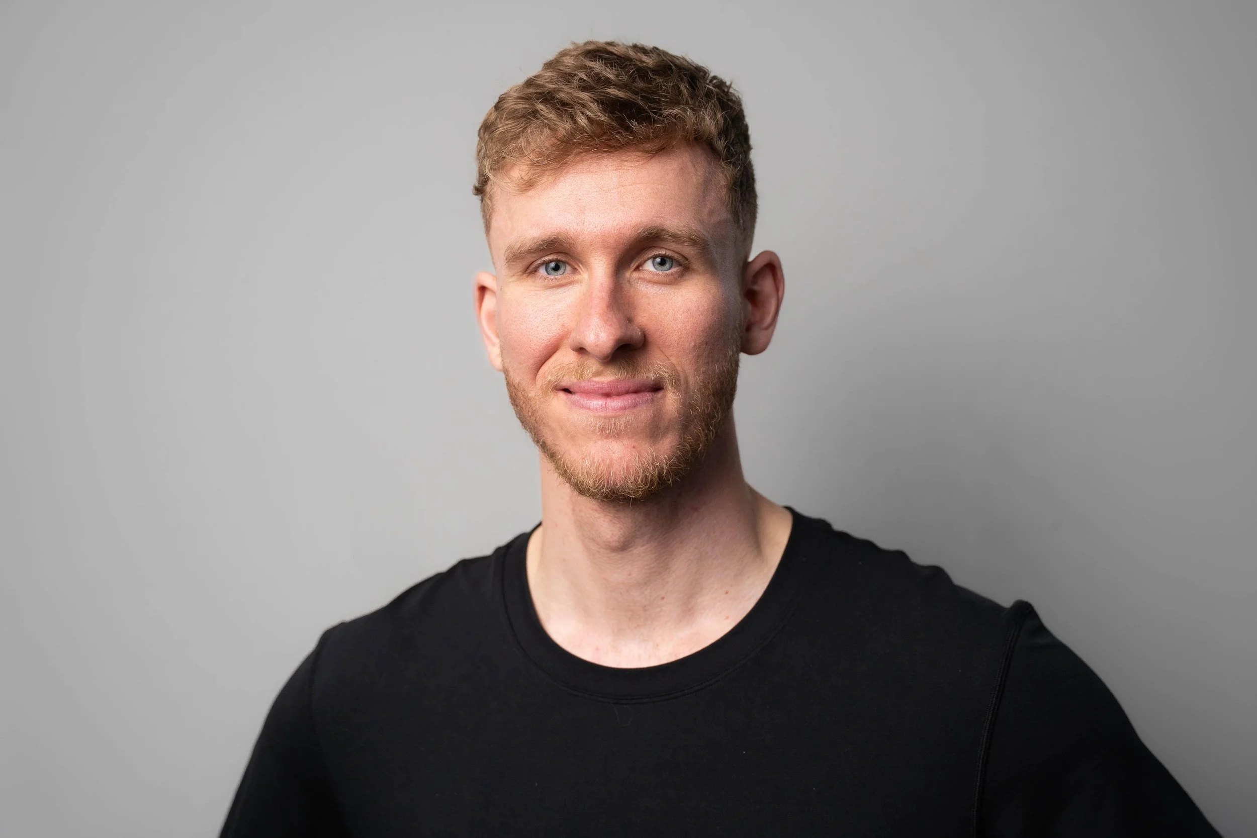Portrait of a young man with short, wavy, light brown hair, blue eyes, and a light beard, wearing a black shirt, standing against a plain gray background.