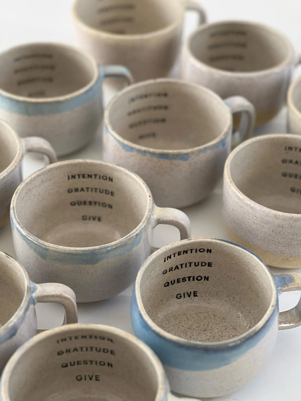 Several ceramic coffee mugs with inner sayings, some with words like 'INTENTION,' 'GRATITUDE,' 'QUESTION,' and 'GIVE' visible inside.