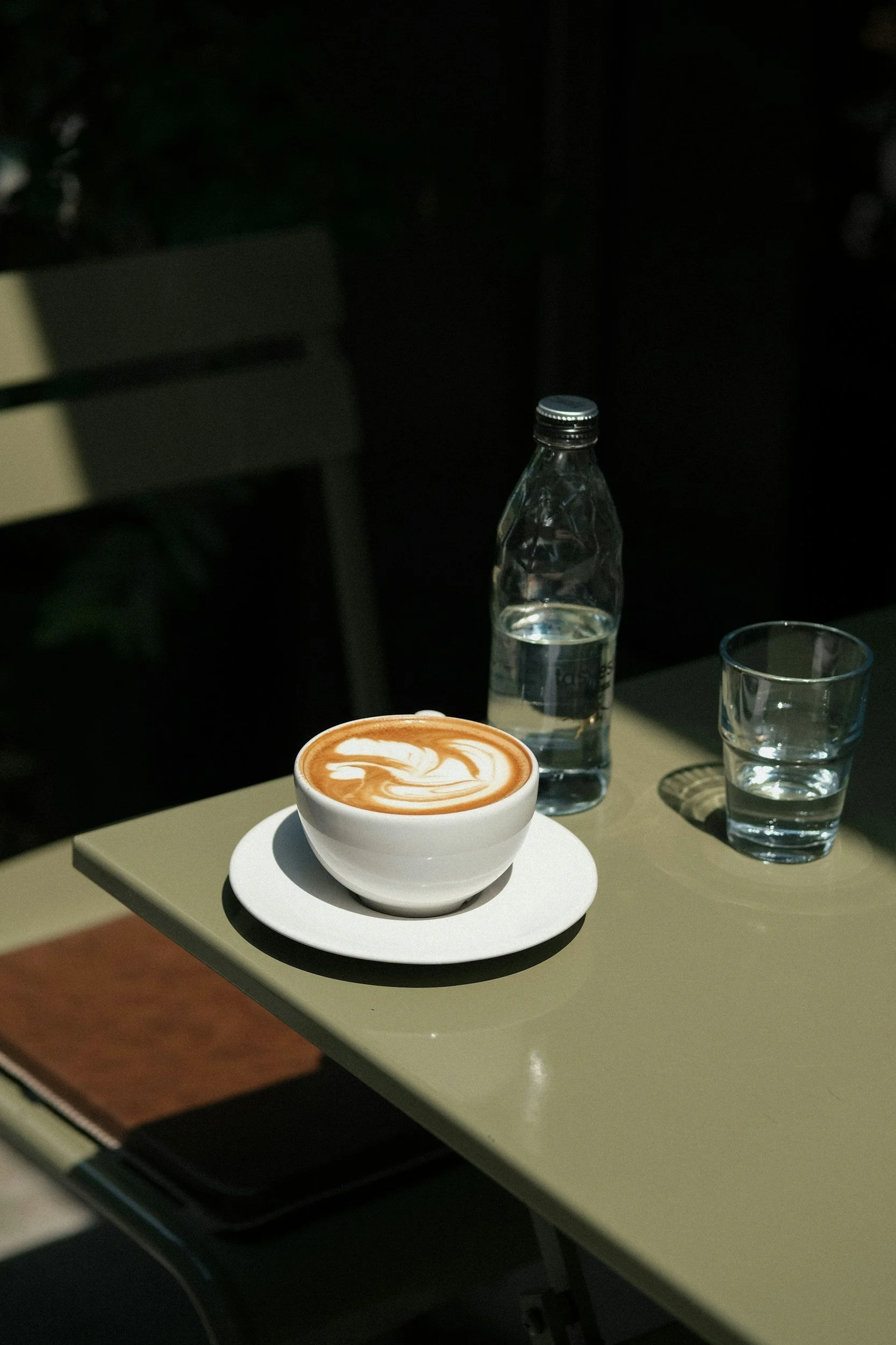 Cup of cappuccino with latte art placed on an outdoor café table next to a glass bottle of water and two empty glasses, in soft natural light – The Family Atelier, Milan.