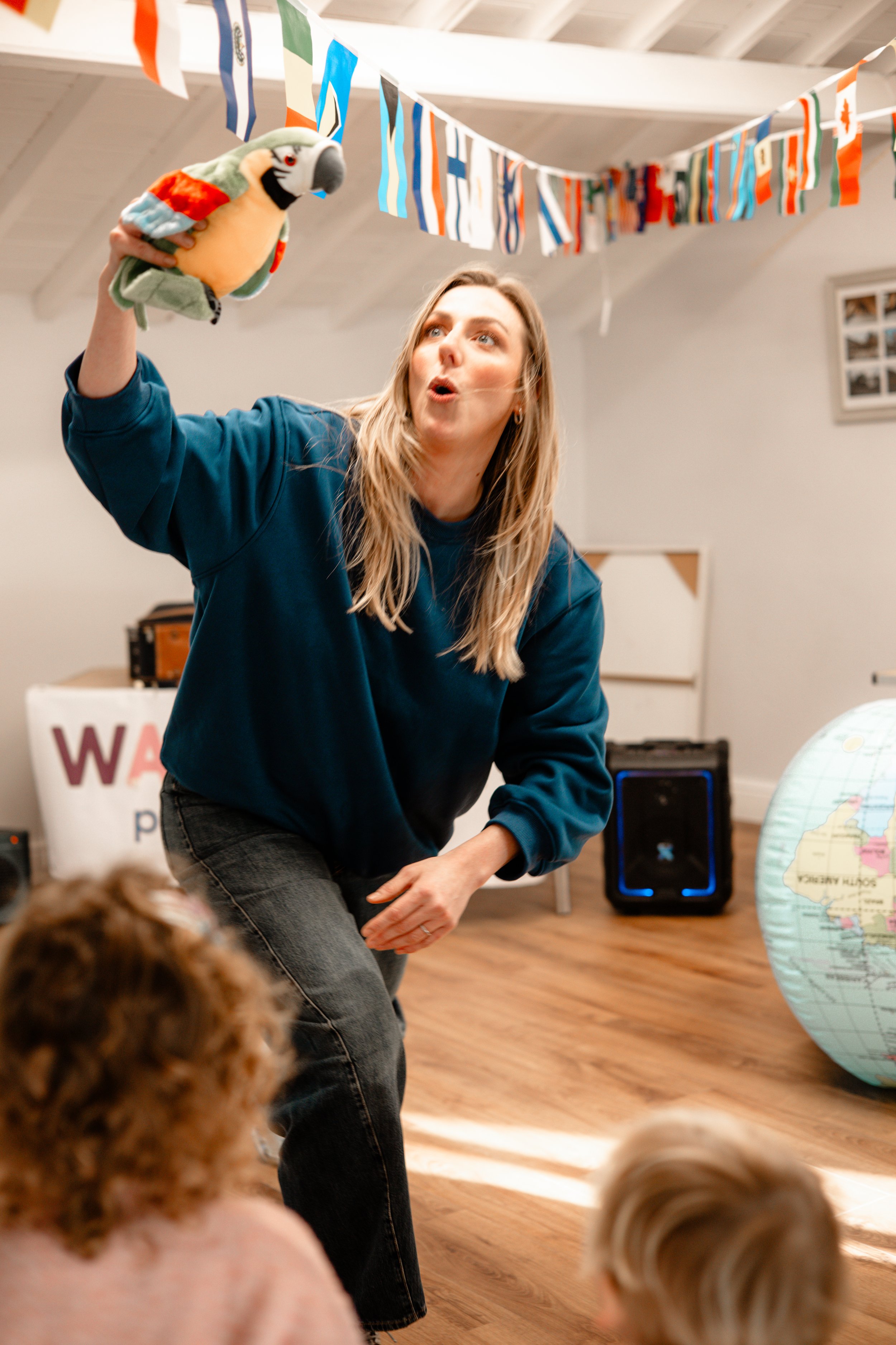 A woman with long blonde hair, wearing a blue sweatshirt, is holding a colorful parrot puppet and gesturing as if she is performing or telling a story to a group of children. The room has international flags hanging from the ceiling, a globe on the right, and a speaker in the background.
