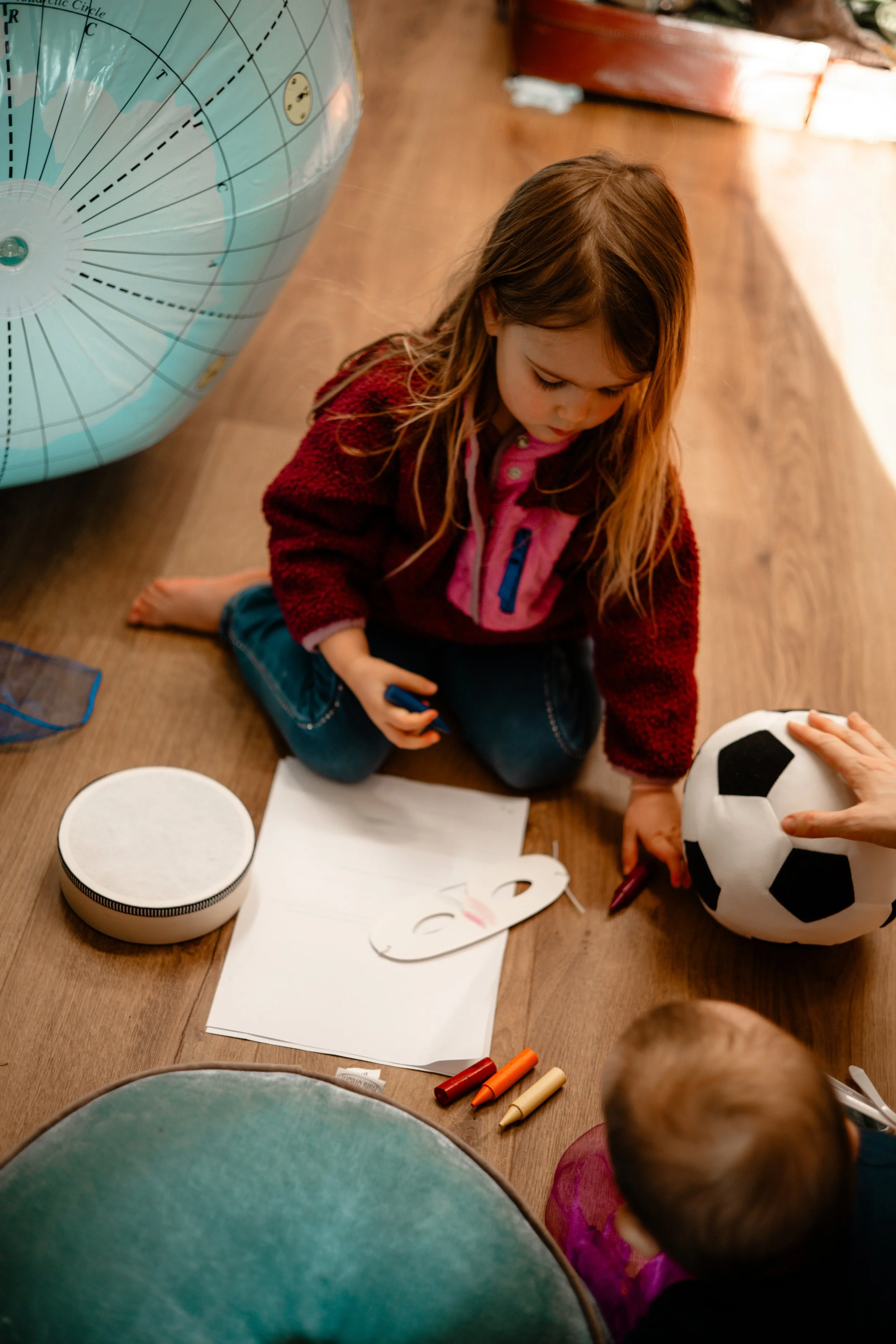 A young girl with long brown hair sitting on a wooden floor, coloring or drawing with markers. There is a soccer ball nearby and a sheet of paper with a face sketch. Part of a large blue globe and some foam rollers are visible in the background.
