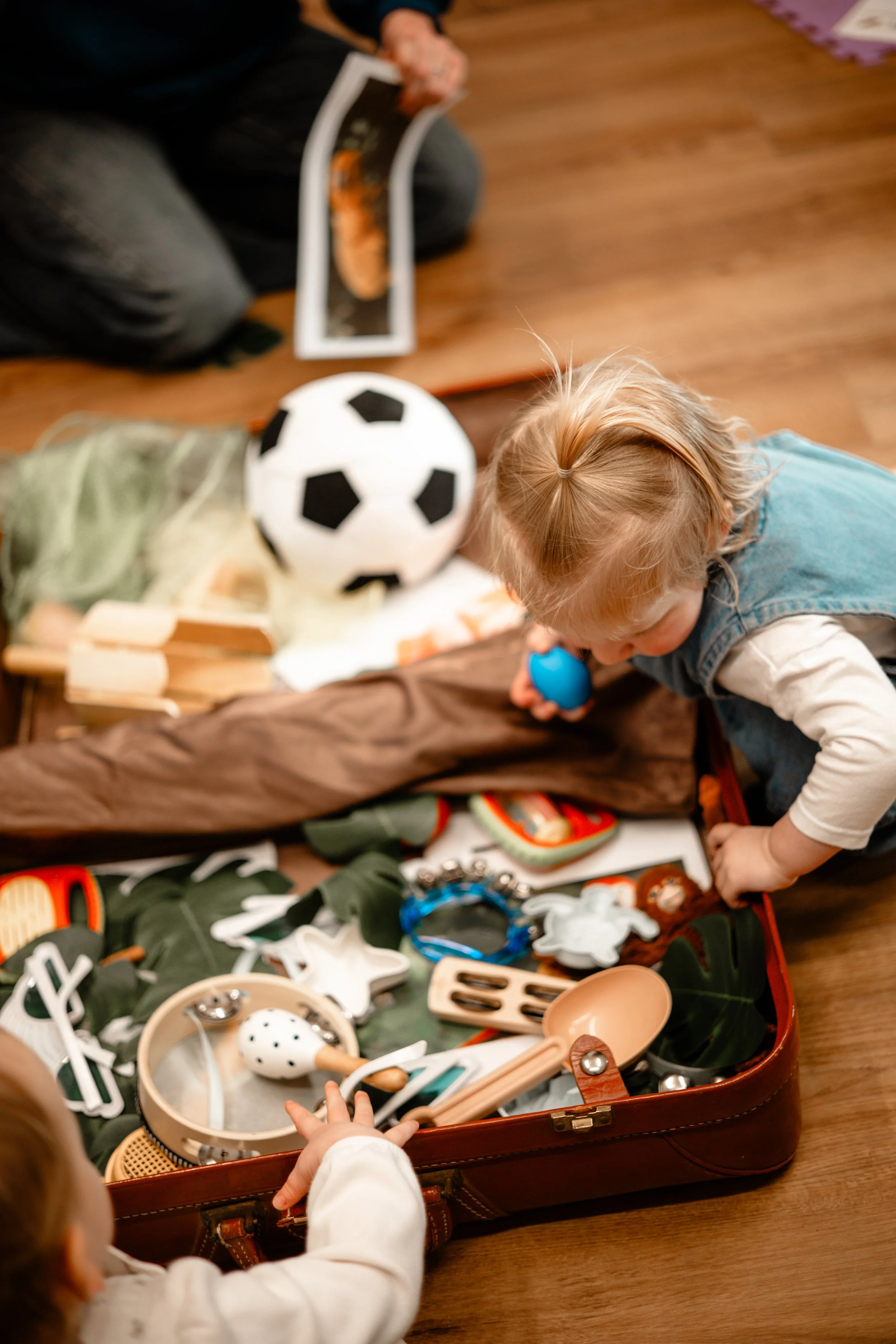 Children playing with toys spilling out of an open suitcase on a wooden floor, including a soccer ball, kitchen utensils, and figurines.