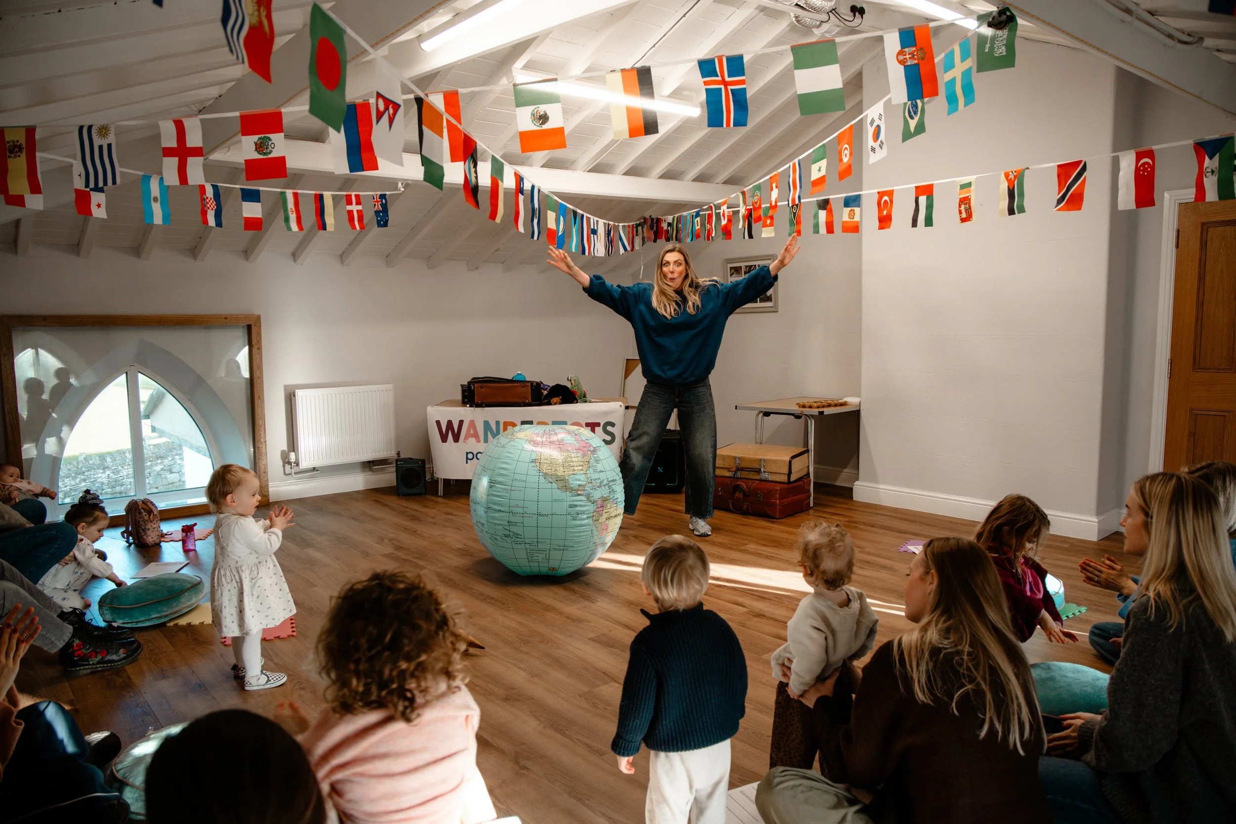 A woman leading a children's educational activity in a room decorated with international flags hanging from the ceiling. The woman is balancing on one foot with her arms outstretched, standing in front of a large inflatable globe. Several children and adults are seated on the floor around her, watching and participating.