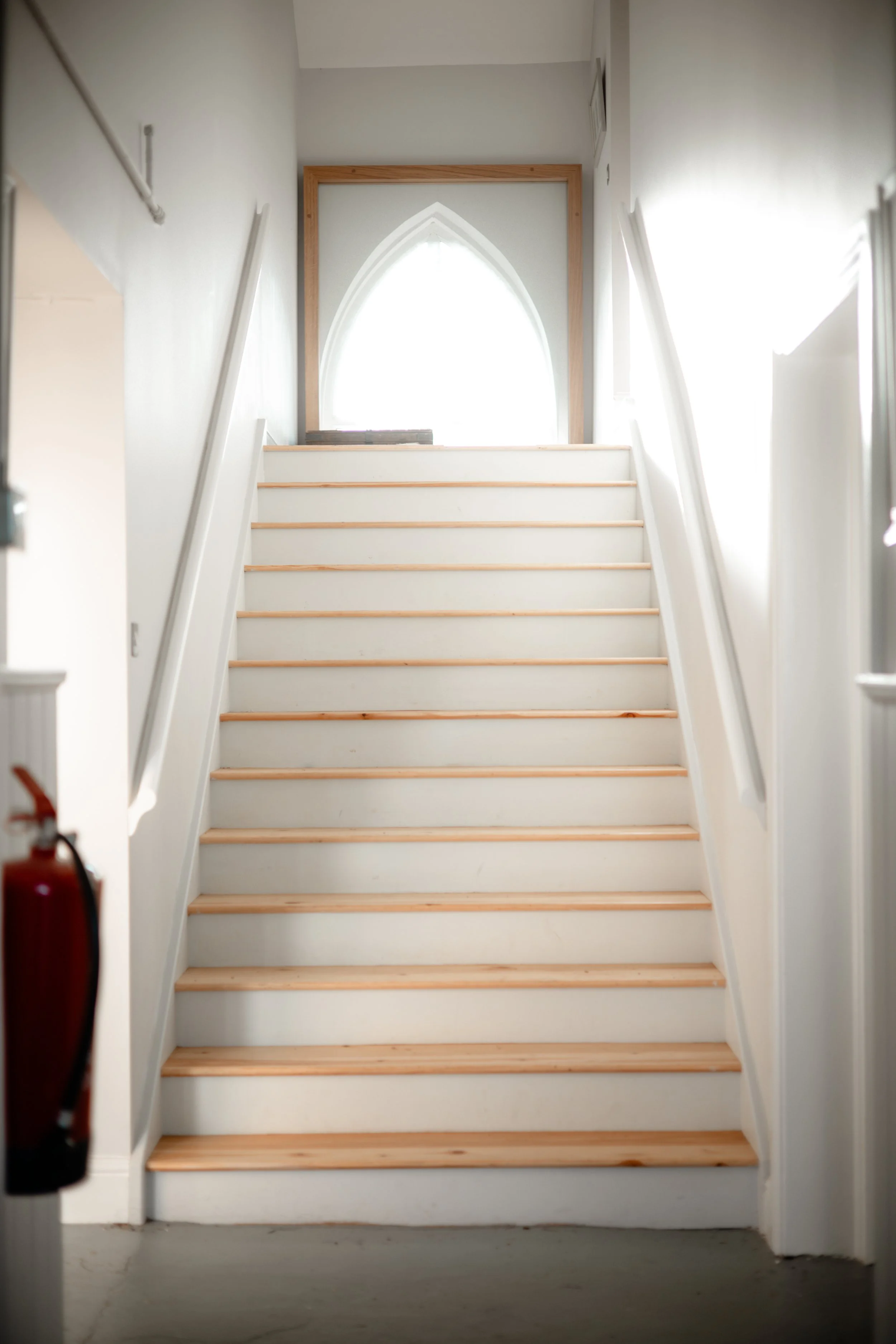 Photo of an indoor staircase with white sides and light wooden steps, leading up to an arched window with bright light shining through, in a building with white walls.