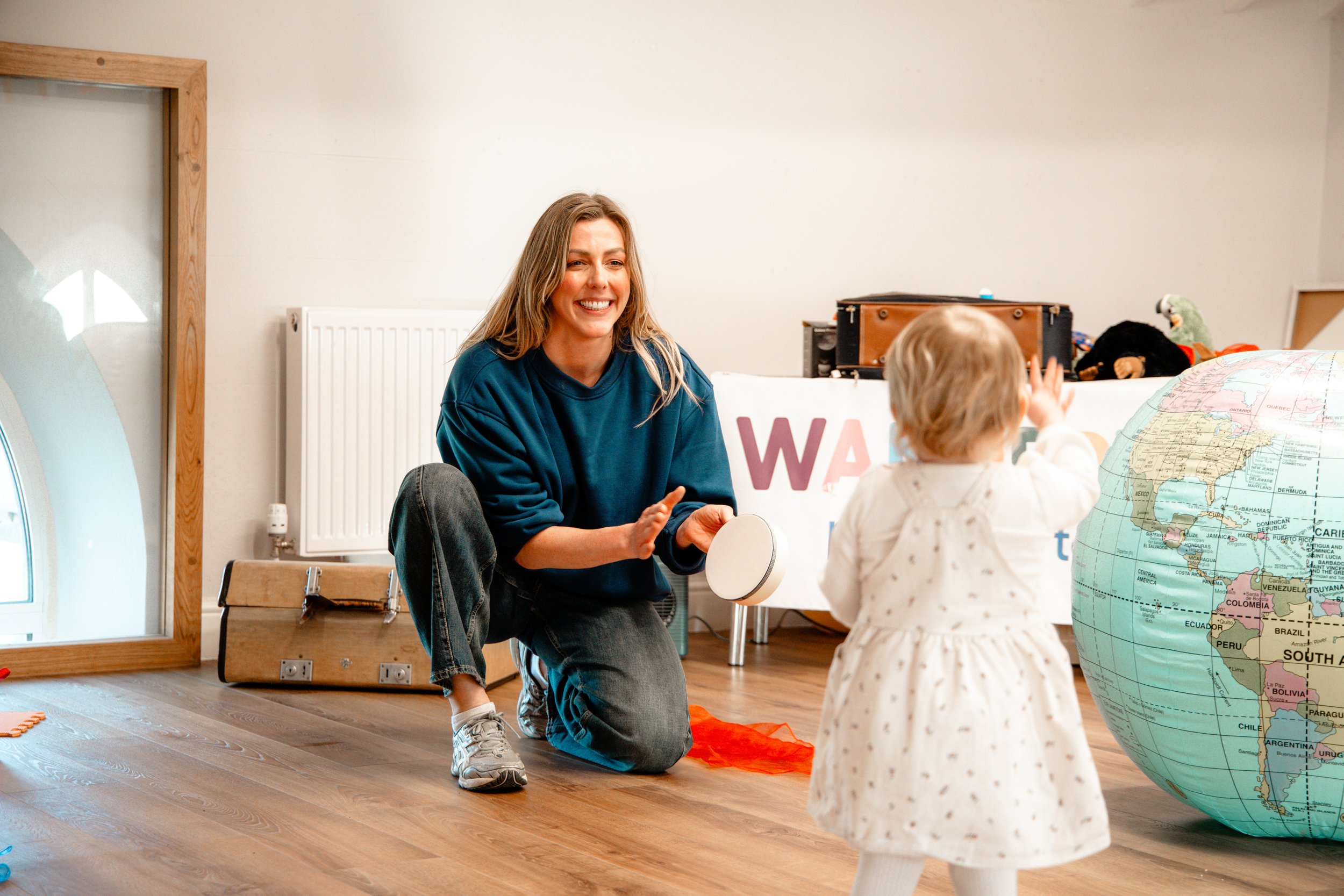 A woman is kneeling on the floor, smiling and playing with a toddler in a room decorated with a world globe, a toy box, and a colorful sign.