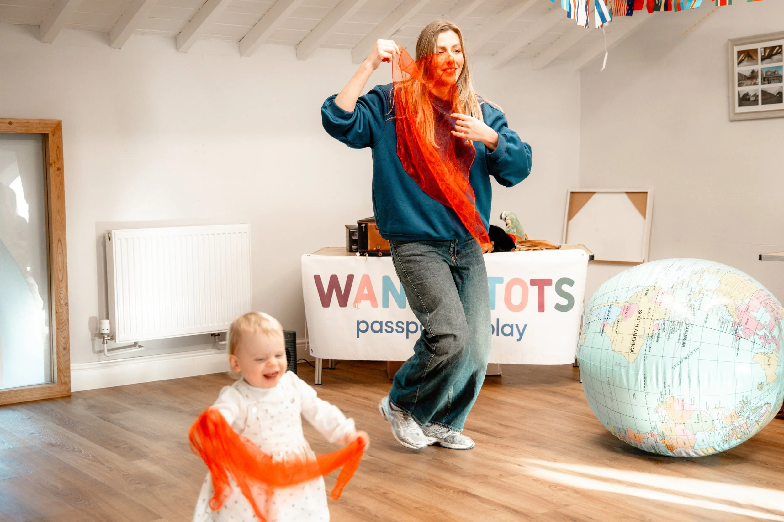 A woman and a young girl playing inside a room decorated for an event. The woman is holding an orange scarf, and the girl is also holding an orange scarf, walking or running happily. In the background, there is a table with a sign that reads 'WANDER TOTS passport holiday,' and a large inflatable globe to the right.