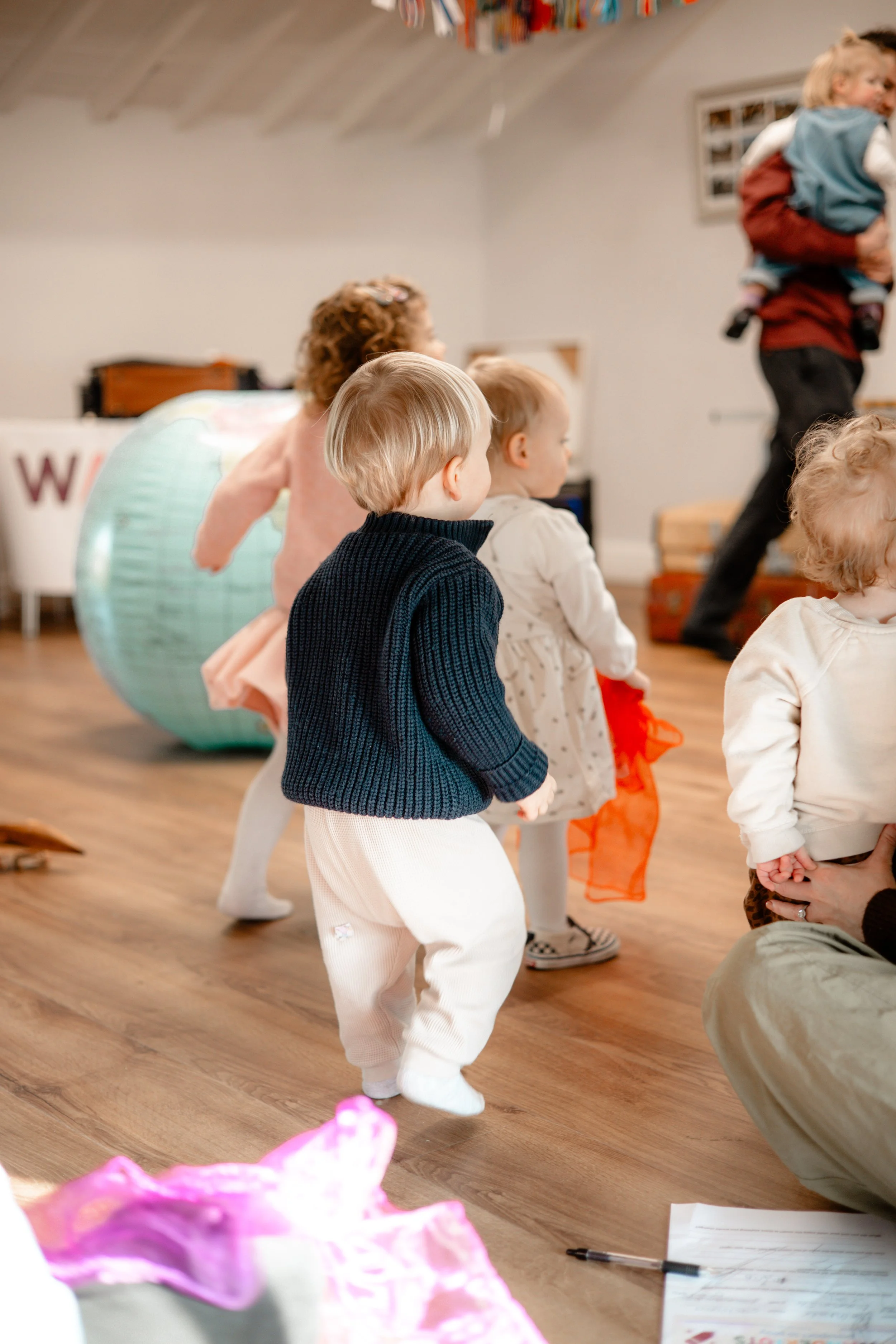 Children participating in a dance or movement activity indoors, with some adults present, on a wooden floor.