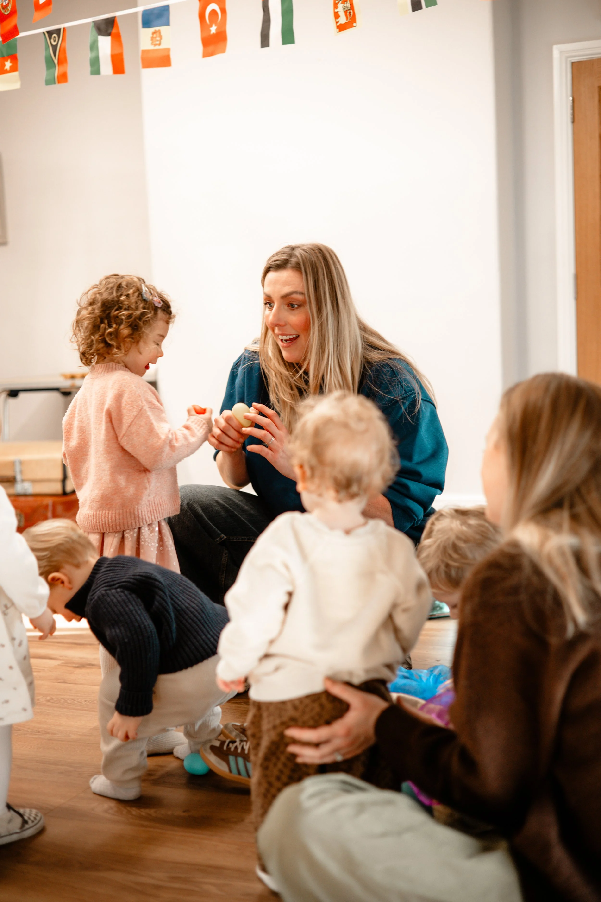 A woman is engaging with children at a party, with some children touching eggs on the floor, with party flags hanging above.