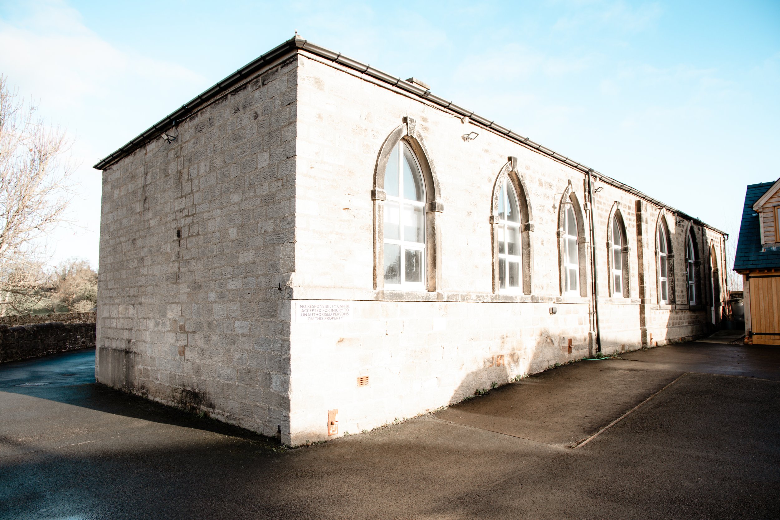 A beige stone building with four tall, narrow arched windows on its side, set against a clear blue sky, with a paved area in front and a wooden structure nearby.