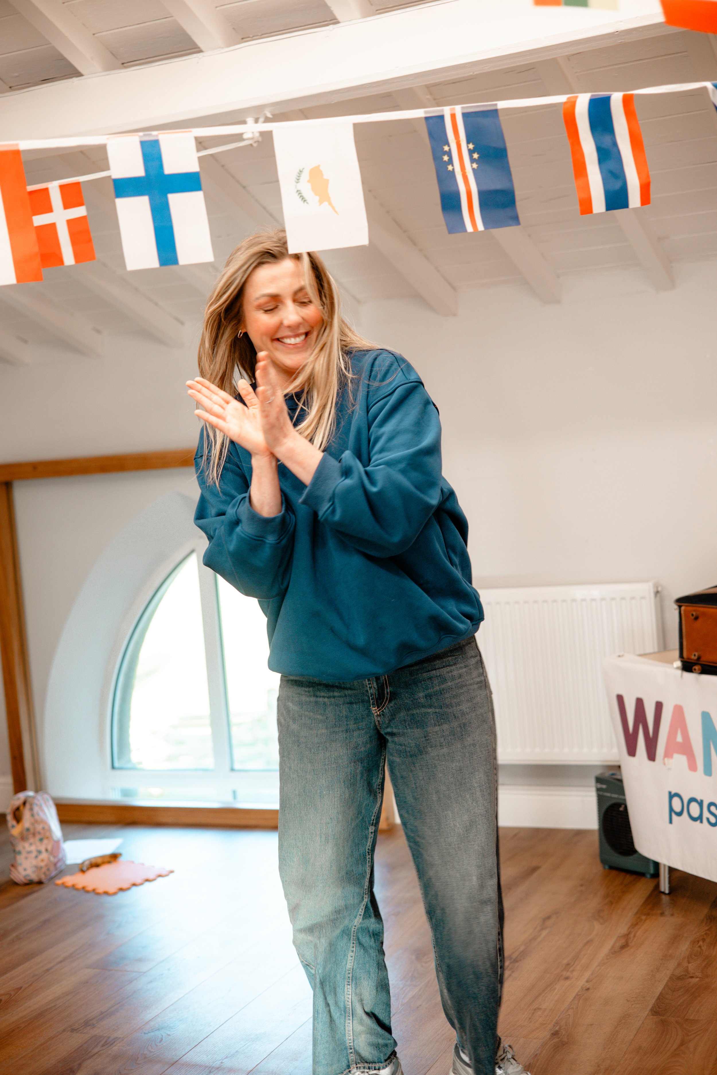 A woman with blonde hair smiling and clapping in a room decorated with international flags.