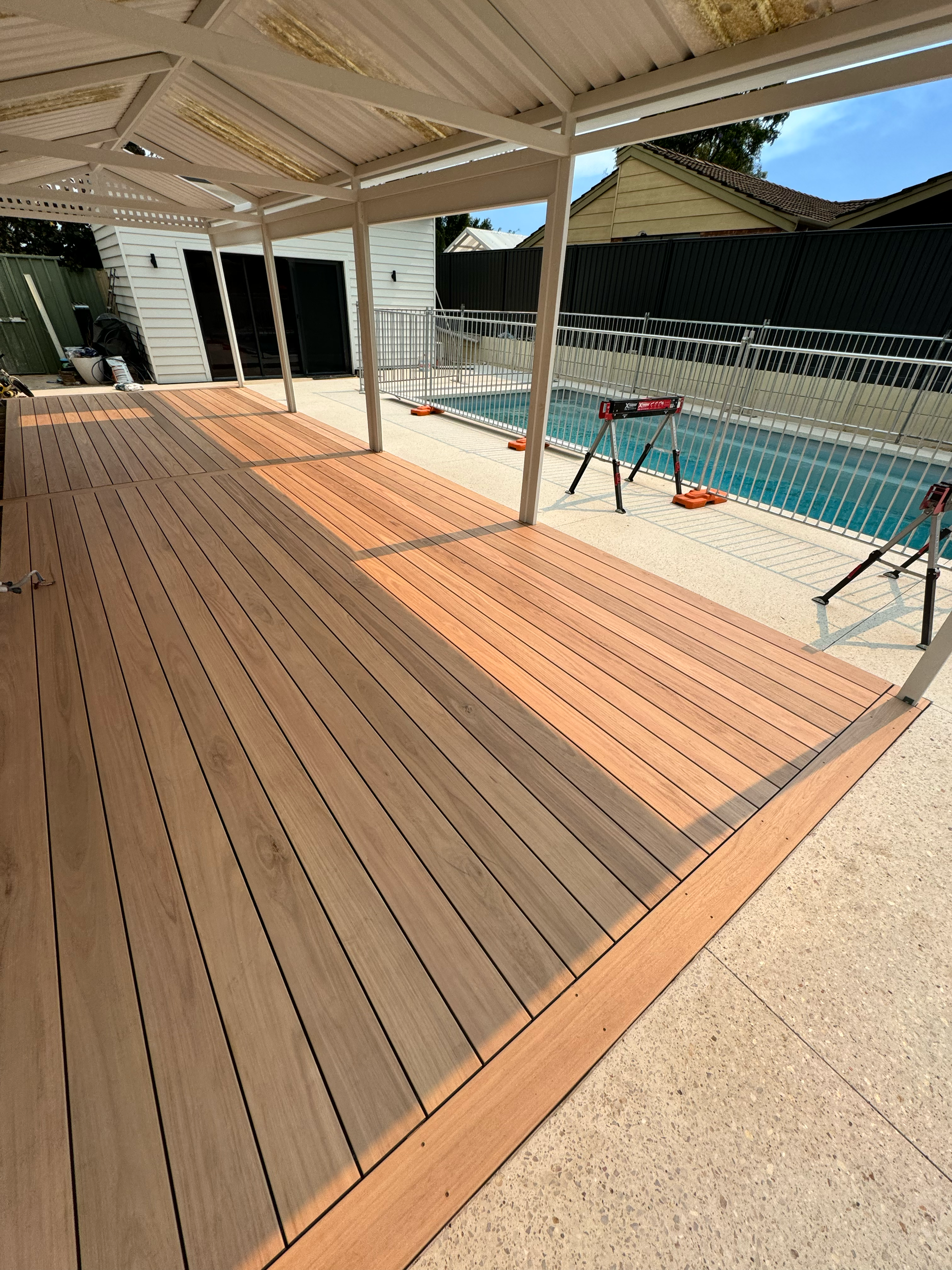 A wooden deck area next to a house with a sliding glass door, with construction tools and materials nearby. A pool and a black fence are visible in the background, along with trees and neighboring houses.