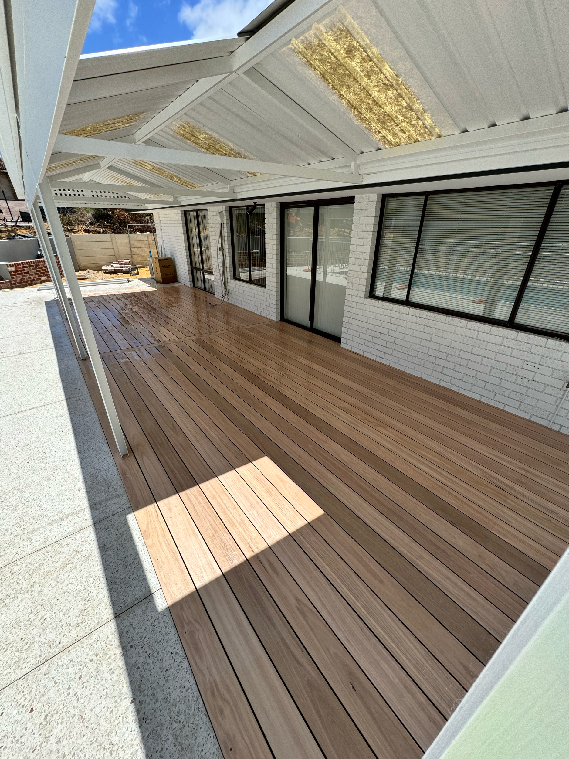 Newly constructed outdoor deck area with wood flooring, enclosed by white brick walls and large windows, under a white metal and glass awning with panels of roofing insulation visible