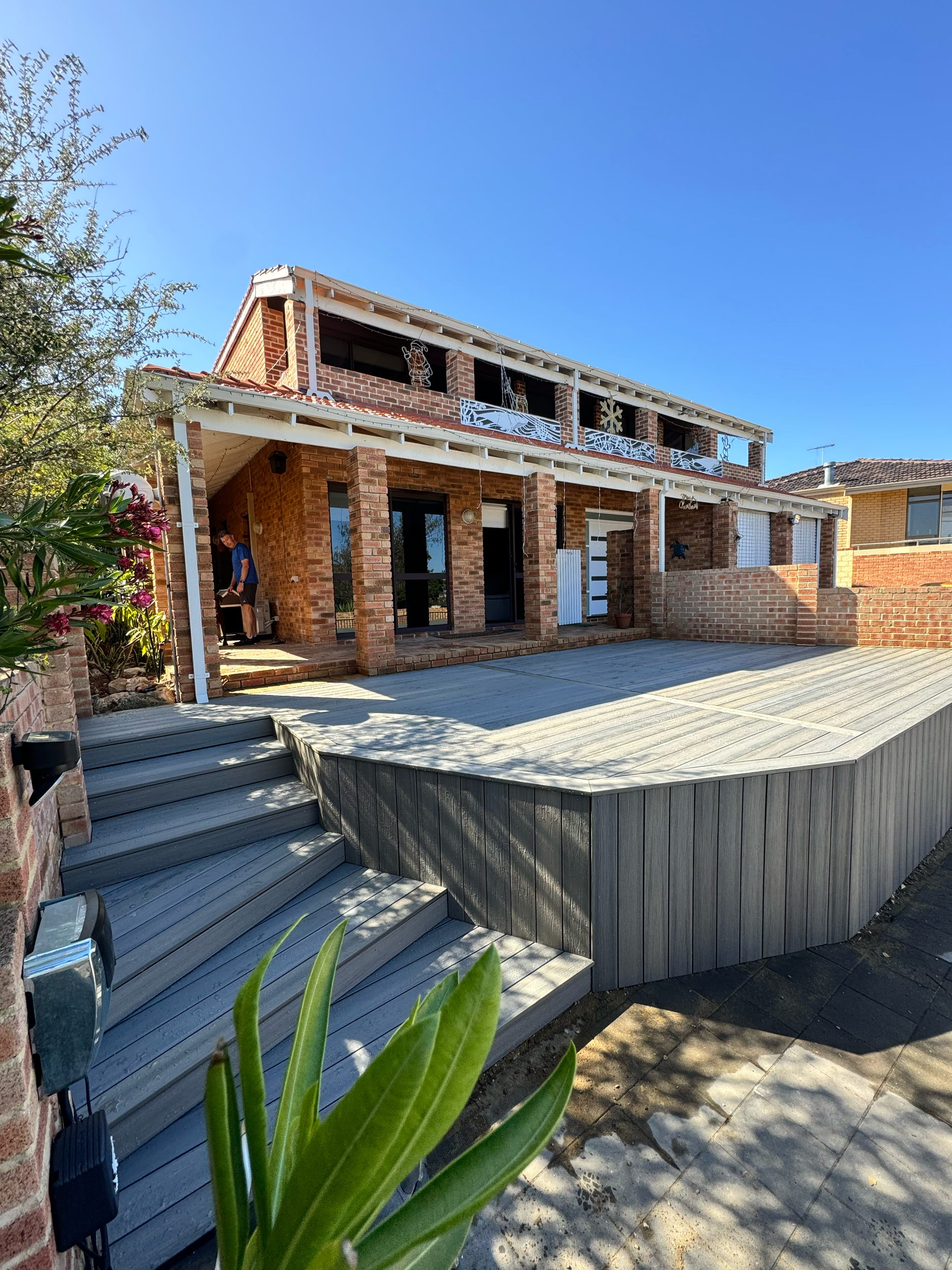 A multi-level brick house with a large wooden deck and balcony, decorated with white railings, surrounded by greenery and set against a clear blue sky.