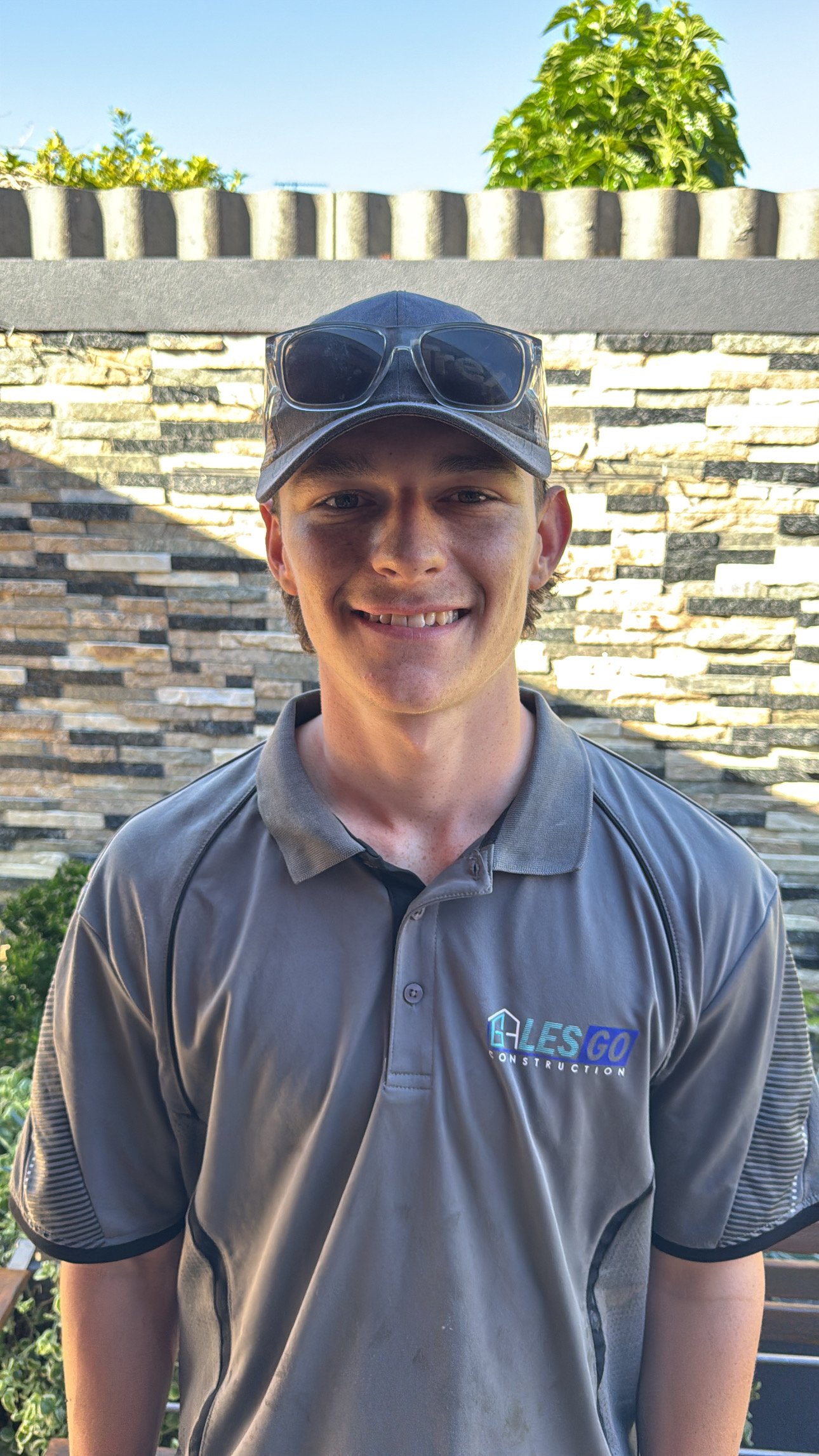 A young man smiling outdoors, wearing a gray polo shirt with a construction company logo, a baseball cap, and sunglasses on top of the cap. Behind him is a brick wall, green bushes, trees, and a clear blue sky.