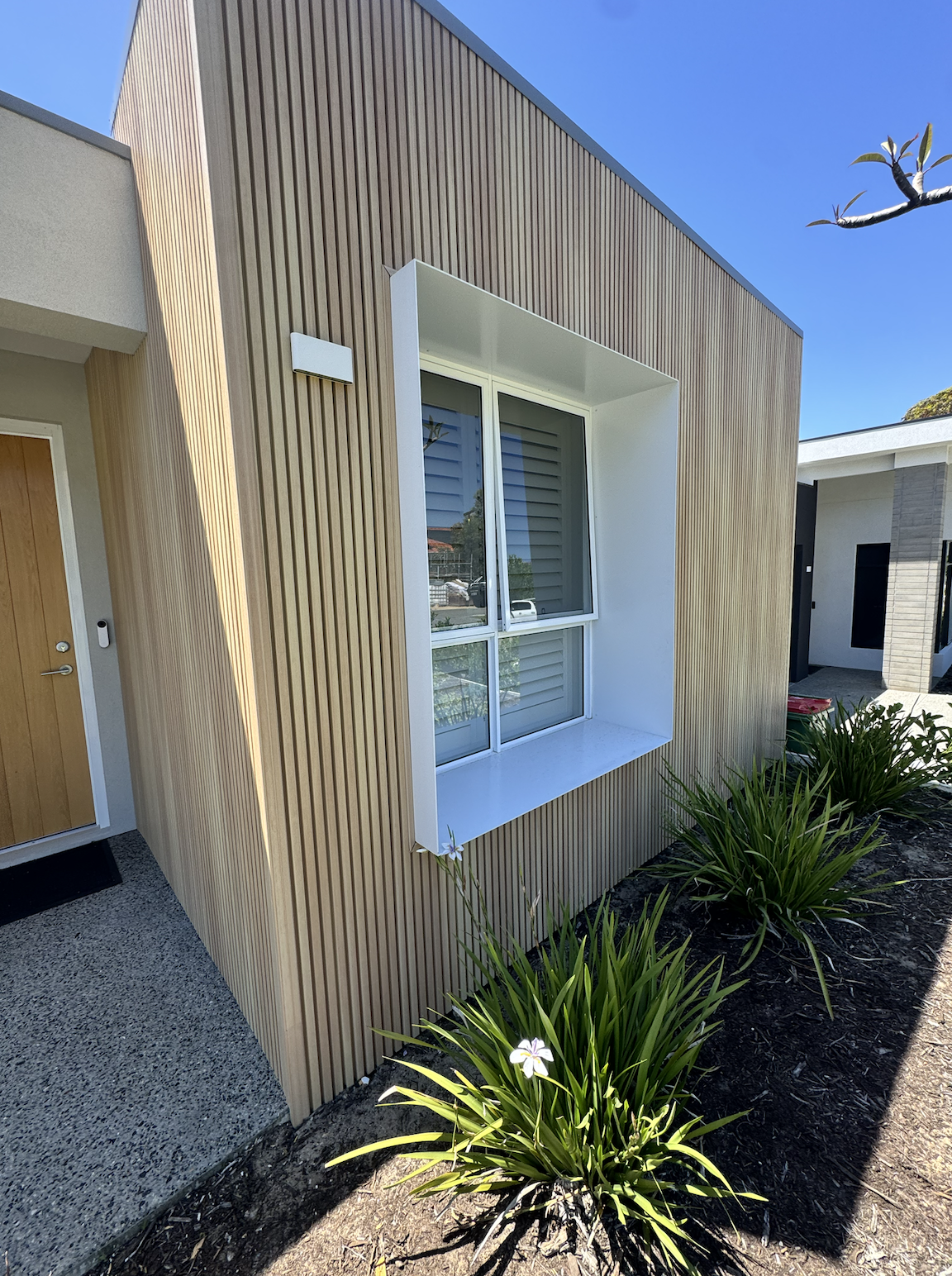 A small outdoor patio with a potted plant, a black wrought iron table, and two wrought iron chairs against wooden cladding.