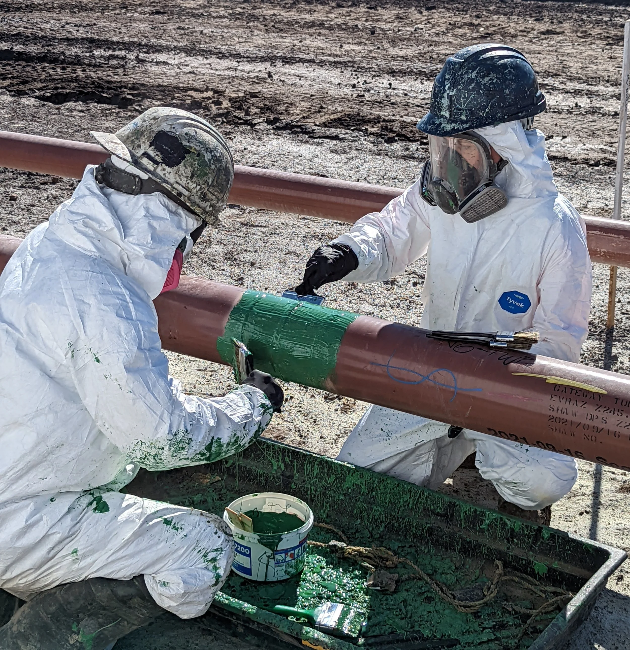 Two workers in protective gear applying green paint or coating to a large metal pipe on an outdoor construction site.