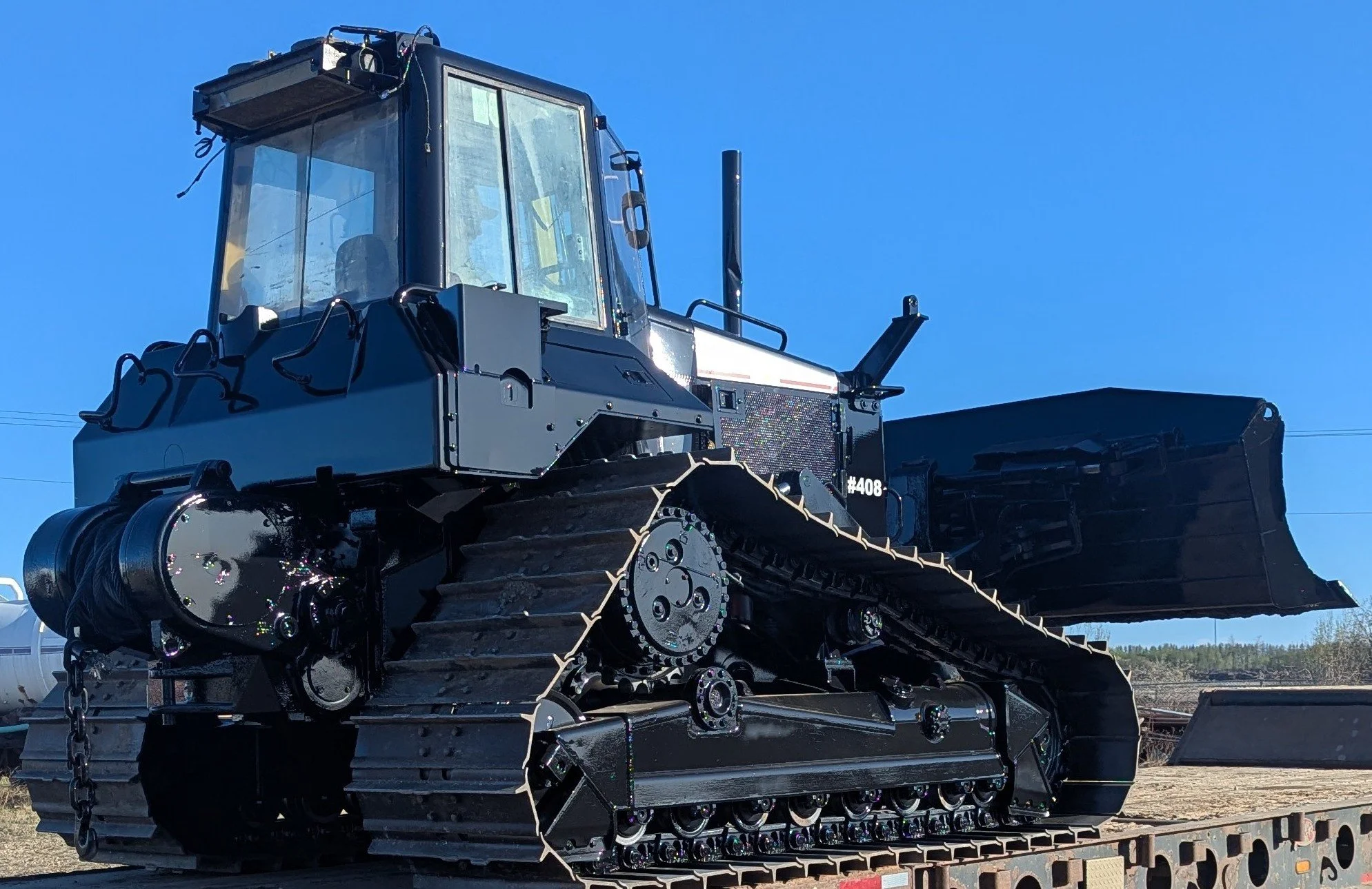 A large black bulldozer with a wide blade at the front, sitting on a flatbed trailer outdoors under a clear blue sky.