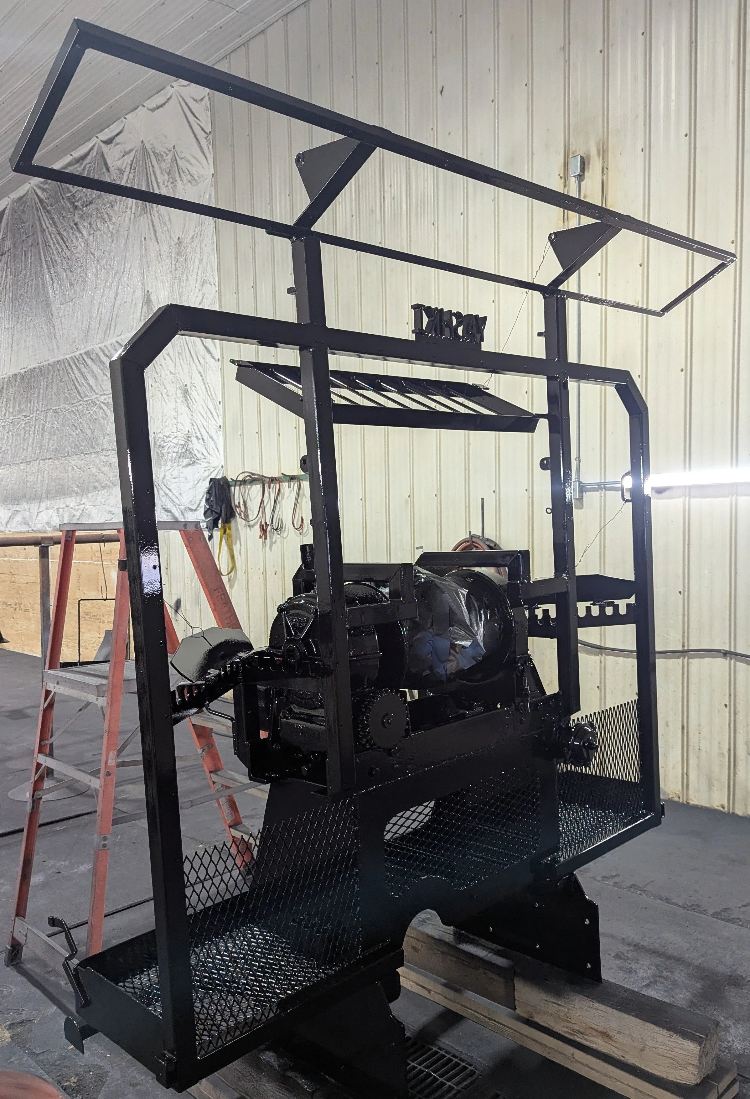 A large black metal frame structure with various mechanical components inside, set inside an industrial workshop with a ladder and tools in the background.