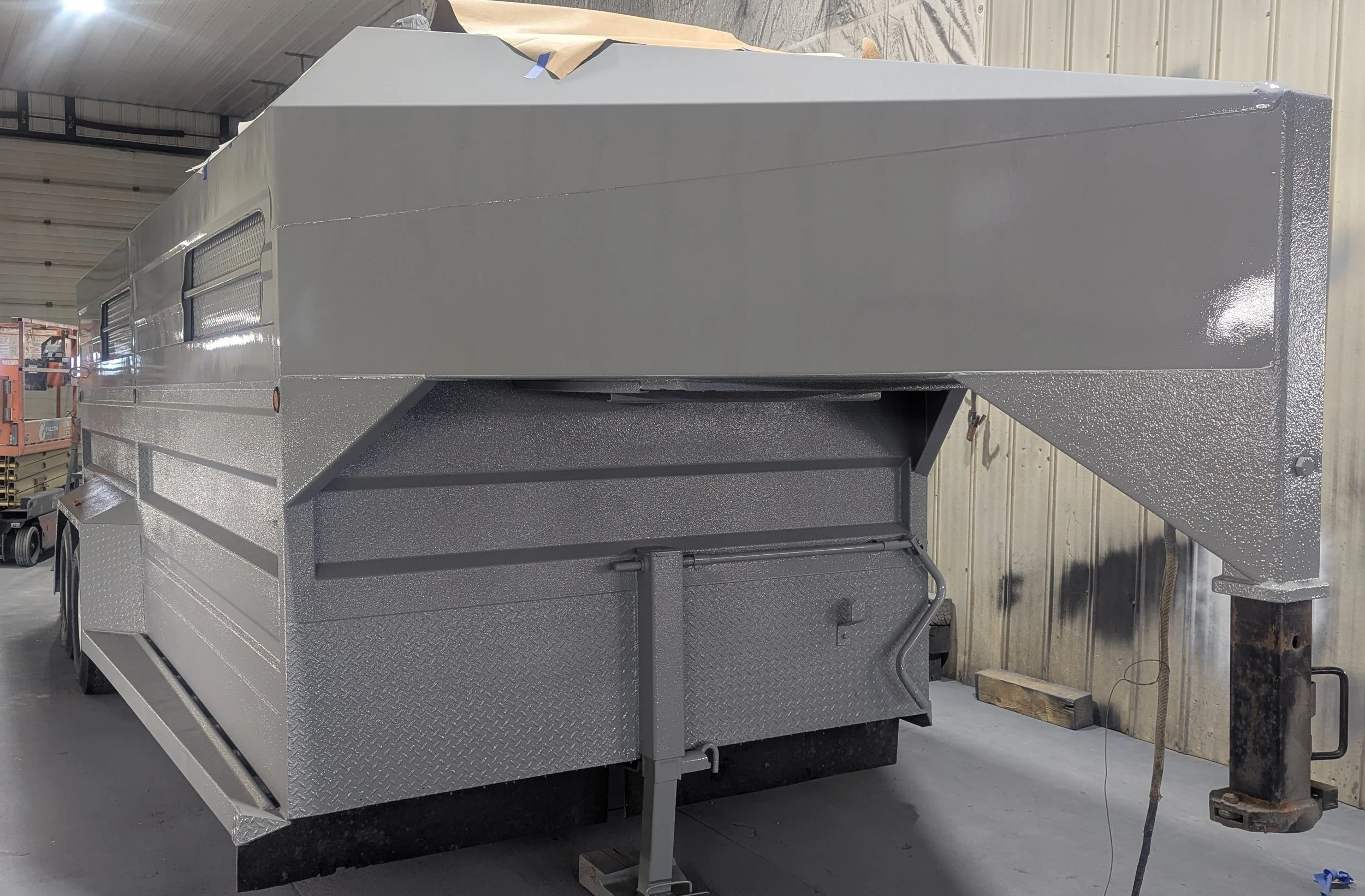 A large, silver metallic trailer or mobile unit inside a warehouse with a high ceiling, with windows along the side, and built-in steps at the side.