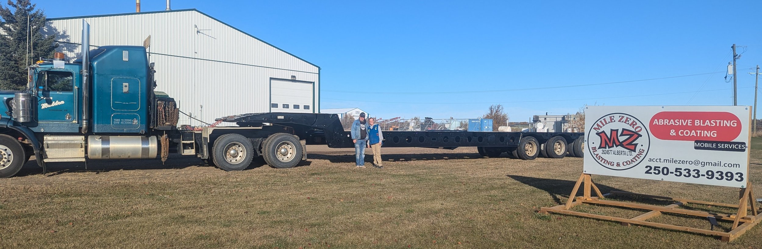 A large blue semi-truck parked on grass near a white industrial building with a sign promoting abrasive blasting and coating services, with two people standing in front of the truck.