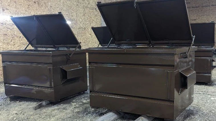 Two large, black metal chest freezers with open lids, situated outdoors on a gravel surface.