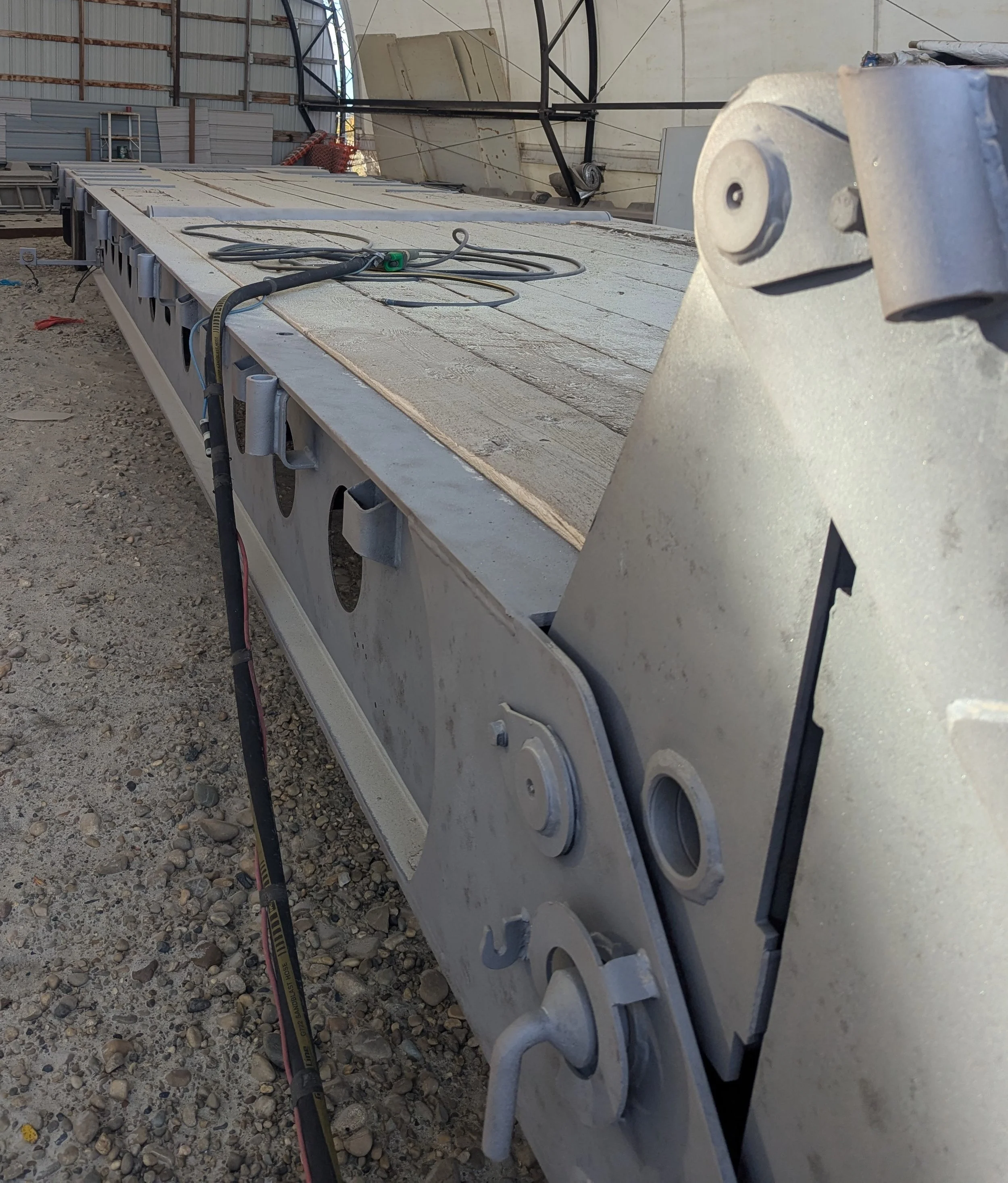 Large flat structure with a metallic base, possibly part of a construction or industrial project, with various cables and tools on top, inside a warehouse or hangar with concrete and metal framework.