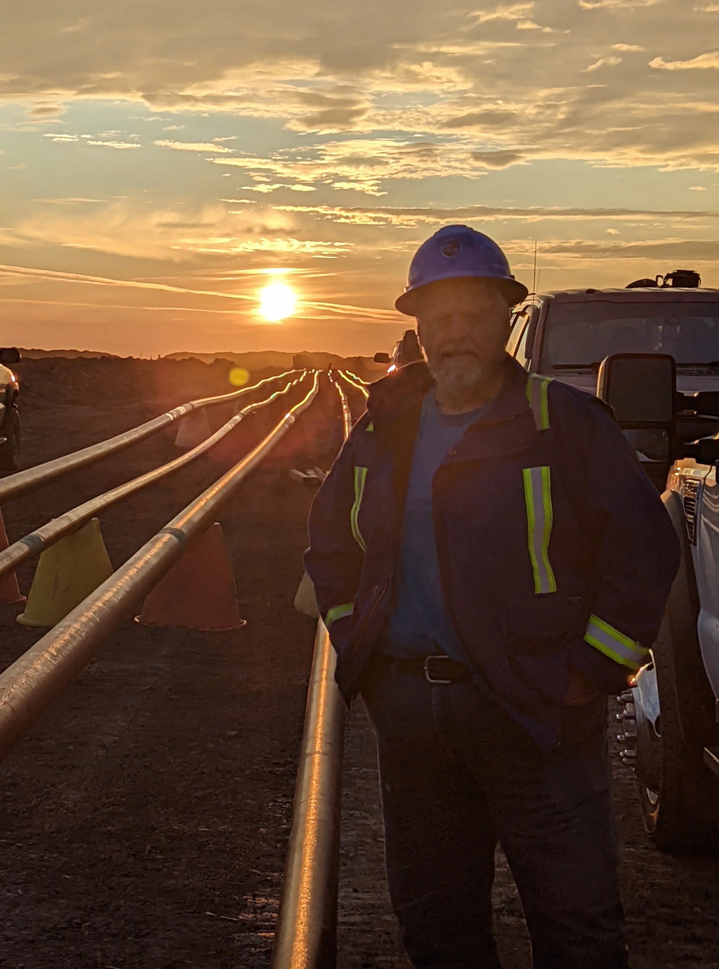 A man wearing a blue hard hat and a dark jacket with reflective strips standing outdoors at sunset, with pipelines and utility vehicles in the background.