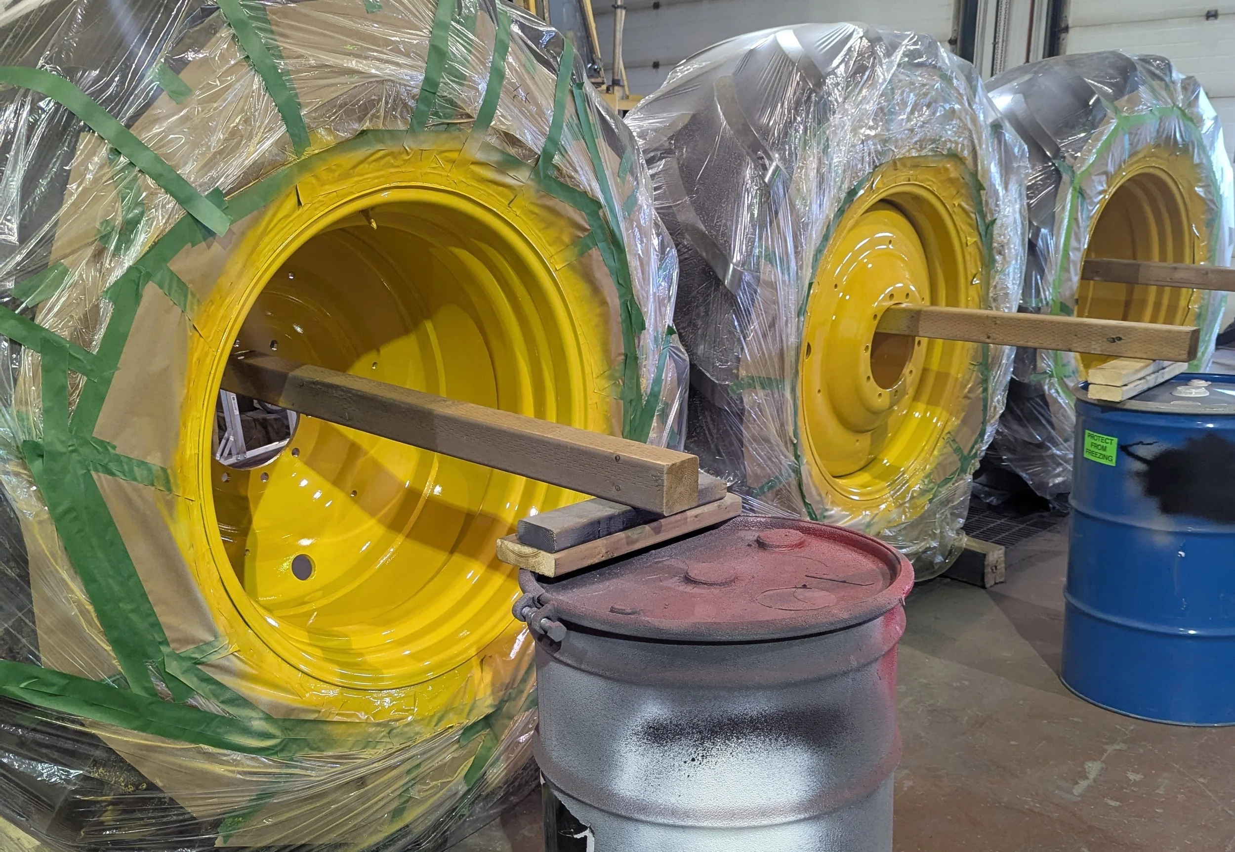 Three large yellow tractor wheels wrapped in plastic and secured with green tape, with a wooden plank through each wheel for support, situated in a workshop or warehouse. There are two barrels nearby, one red and one blue, with the blue barrel having a green label that reads 'Protect from Freezing'.