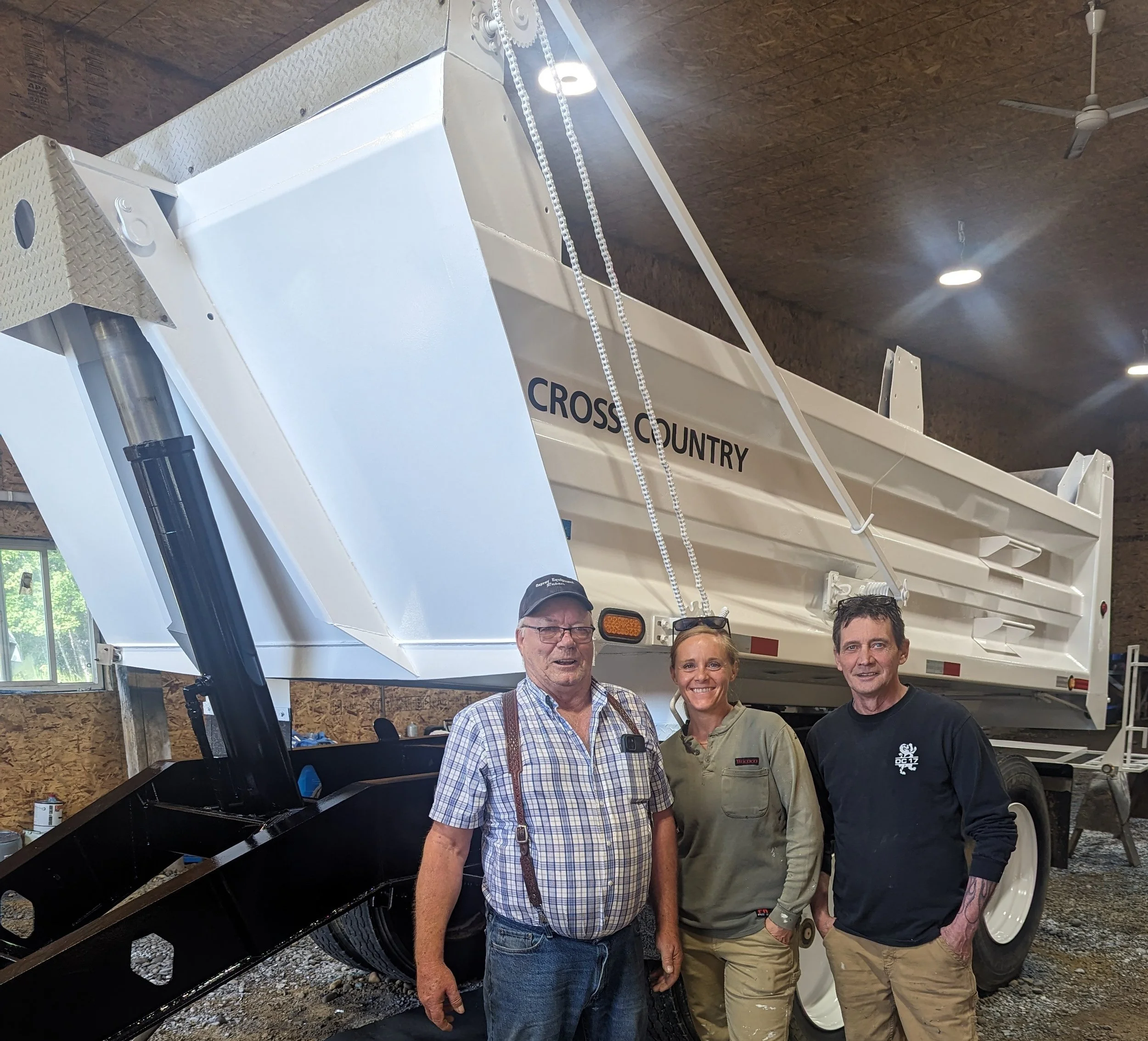 Three people standing in front of a large, white Cross Country grain cart inside a barn or workshop.