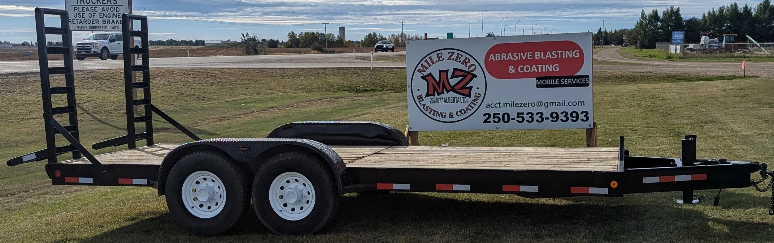 A flatbed trailer with dual tires parked on grass, with a sign in the background advertising Mile Zero Blasting & Coating, a mobile abrasive blasting and coating service company, including the contact email and phone number. The scene is set near a highway with vehicles passing by.