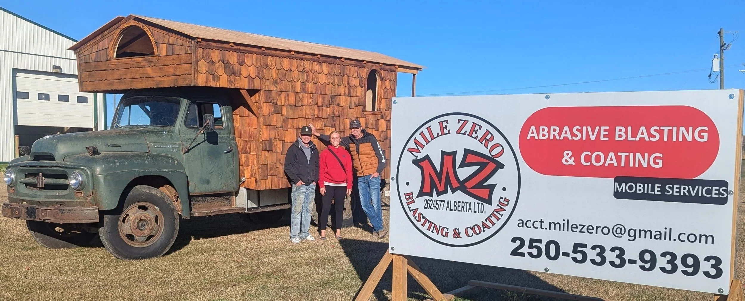 Three people standing next to a vintage truck with a wooden structure on its back, parked beside a sign for Mile Zero Blasting & Coating, advertising abrasive blasting and coating services, with contact information and email address.