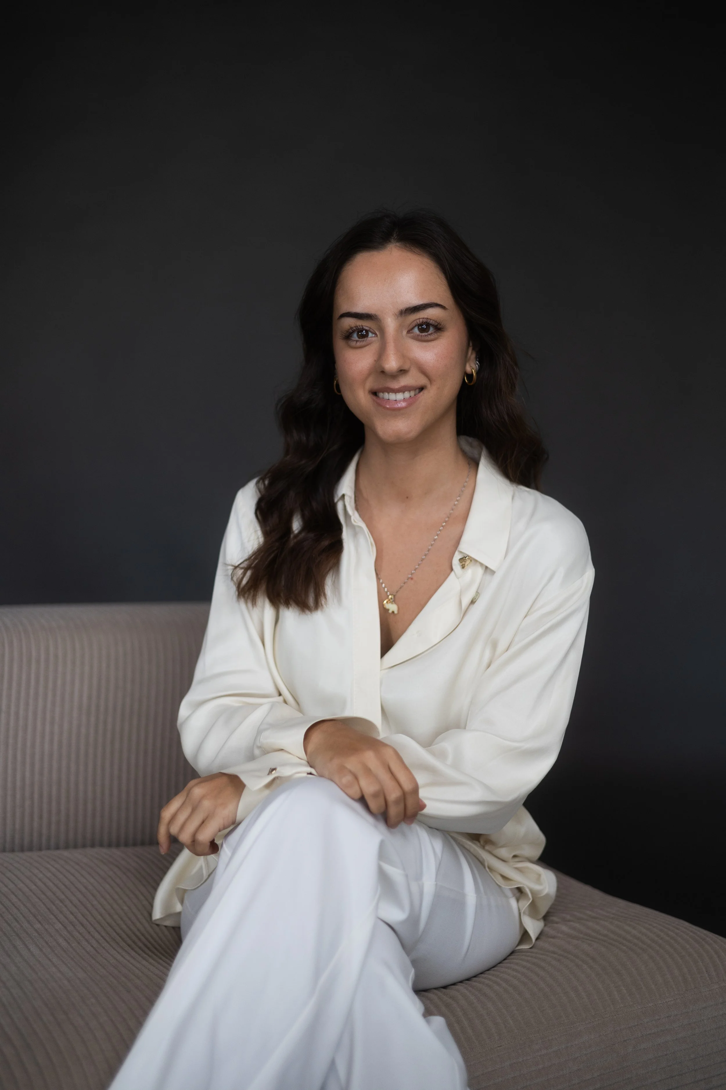 A young woman with dark wavy hair and earrings, sitting on a beige couch in front of a dark gray background, wearing a cream-colored blouse and pants, smiling at the camera.