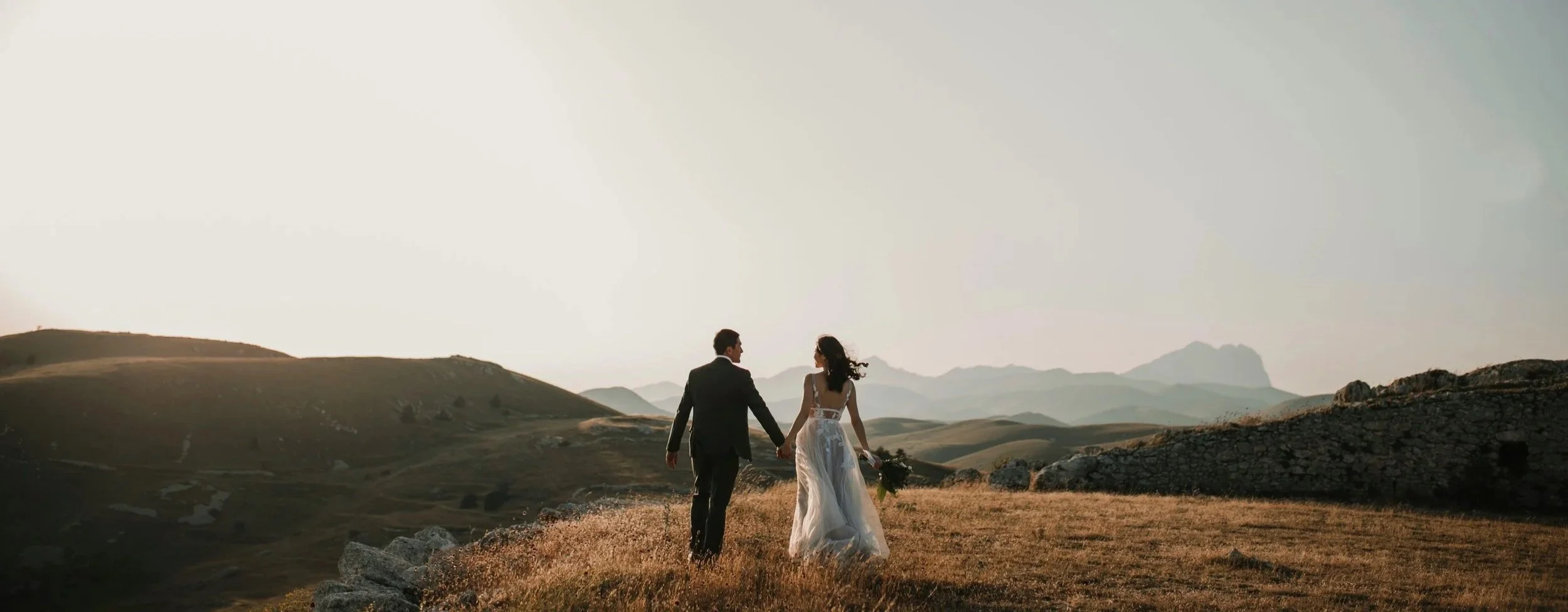 A bride and groom holding hands walking through a grassy field with mountains in the background, during sunset.
