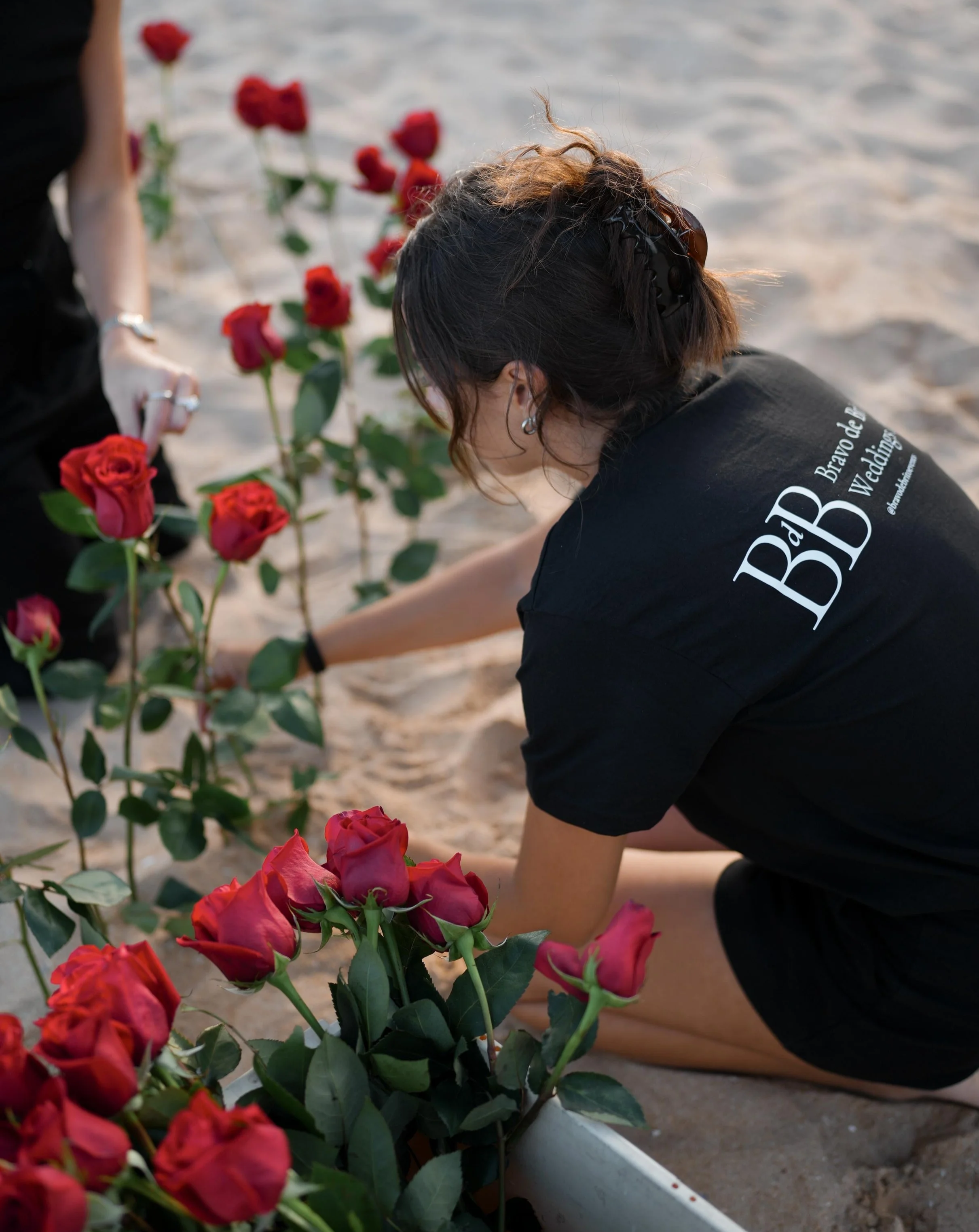 A woman arranging red roses on the sand beach with another person nearby. The woman is wearing a black t-shirt with white text on the back.