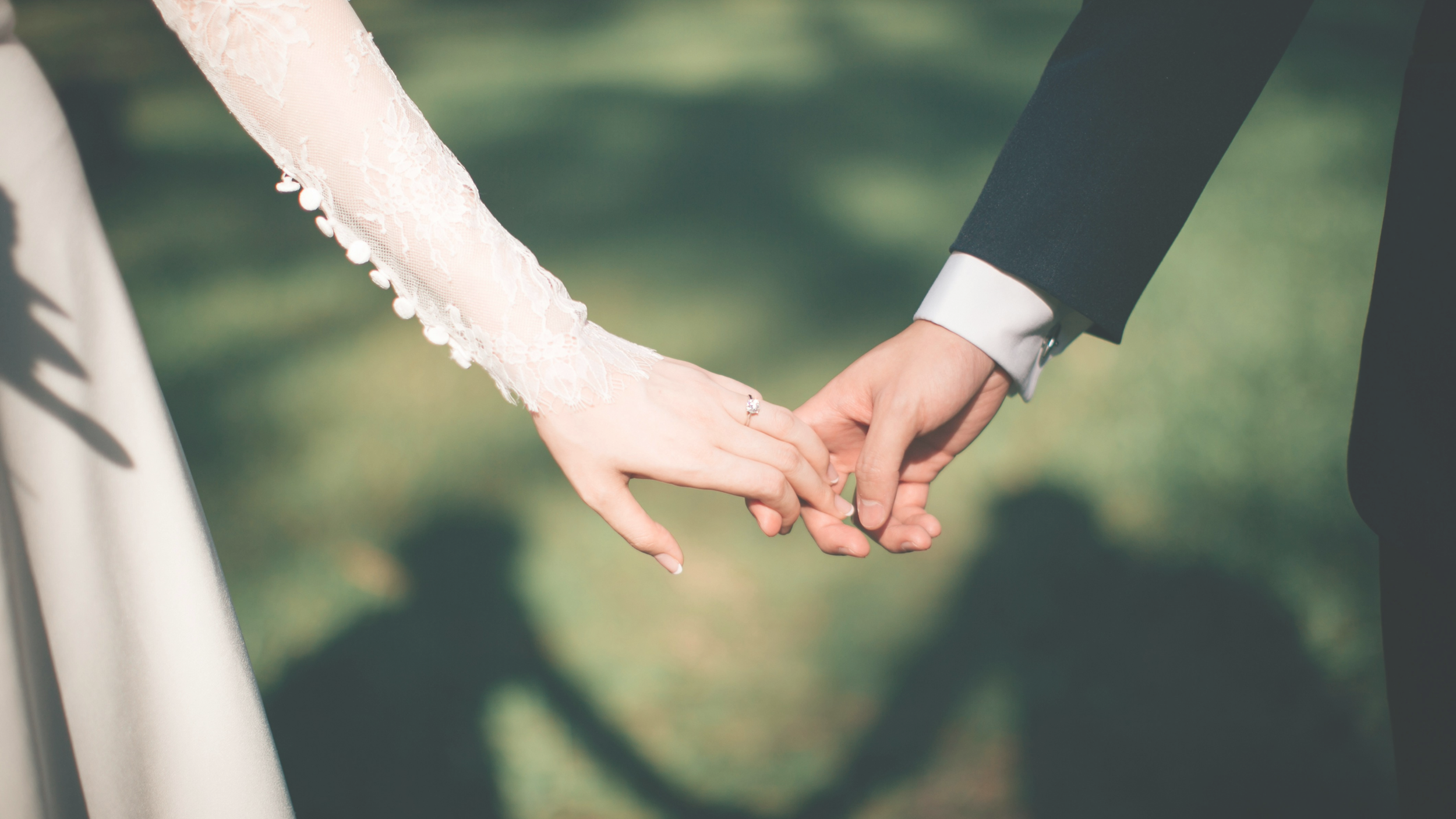 Close-up of a bride and groom holding hands during a wedding ceremony outdoors. The bride wears a lace dress with buttons on her sleeve, and the groom wears a black suit with a white shirt.