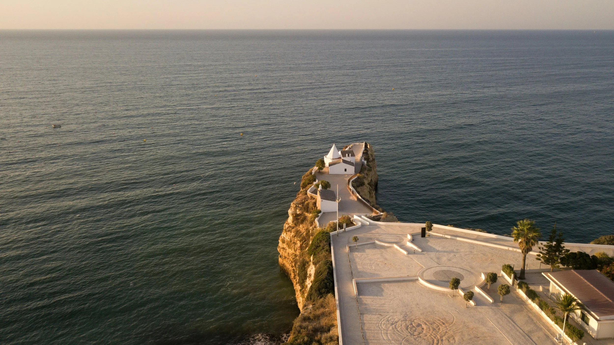 Aerial view of a small white church on a rocky outcrop surrounded by the ocean, with a paved courtyard and palm trees nearby.