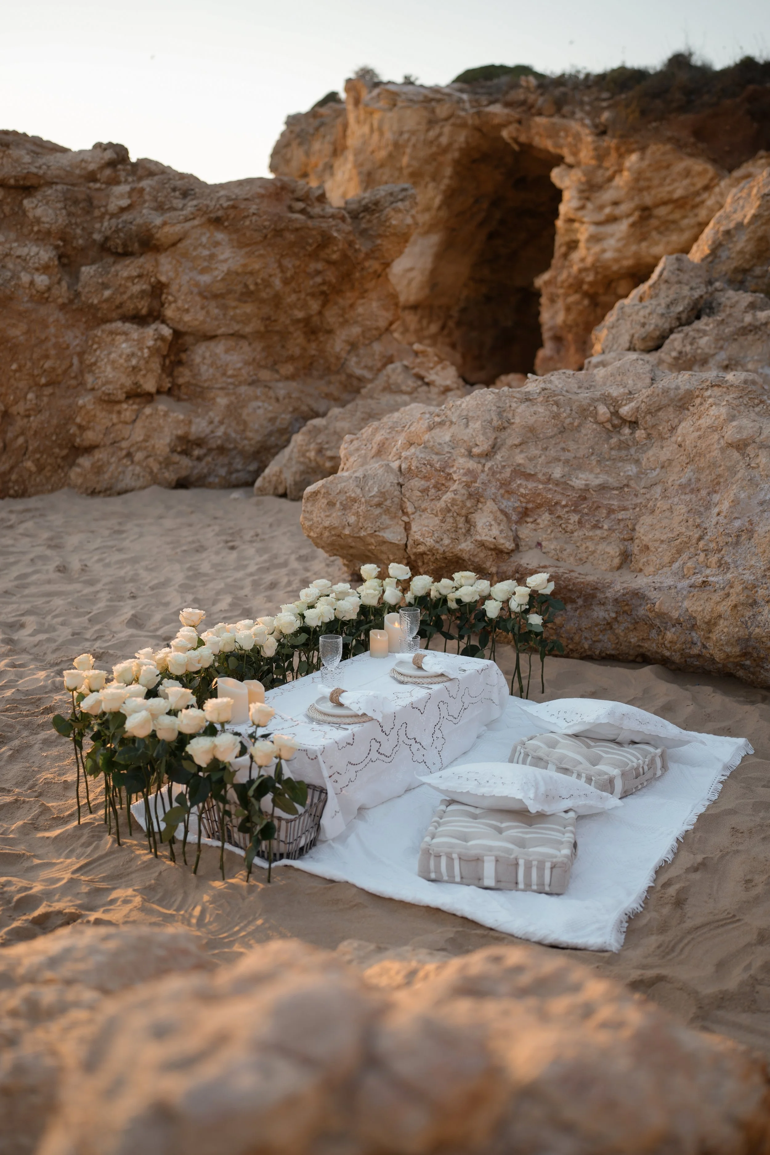 A romantic picnic setup with a low table surrounded by white roses on a sandy beach with large rock formations in the background.