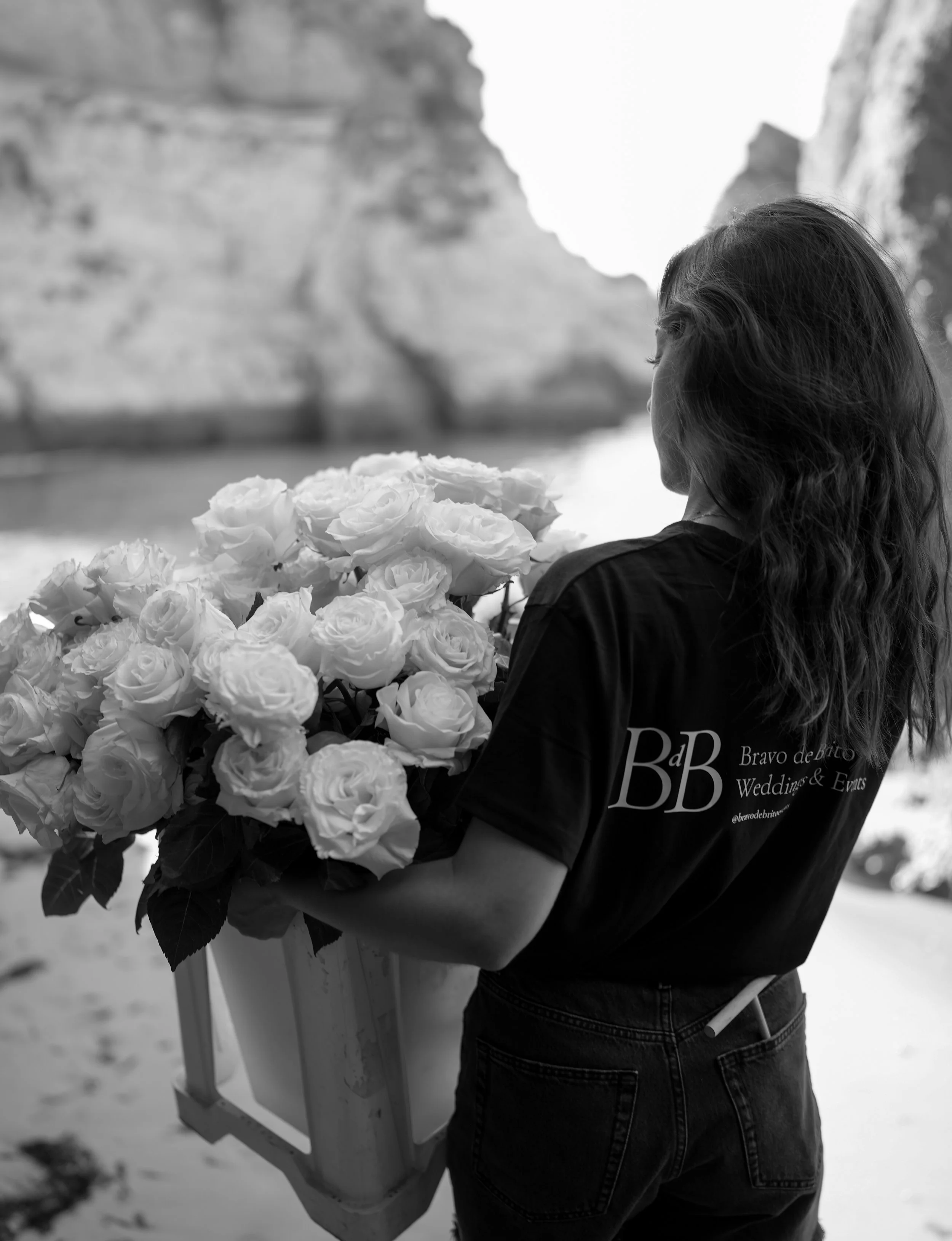A woman with wavy hair holds a large bouquet of roses on a beach, with cliffs in the background.