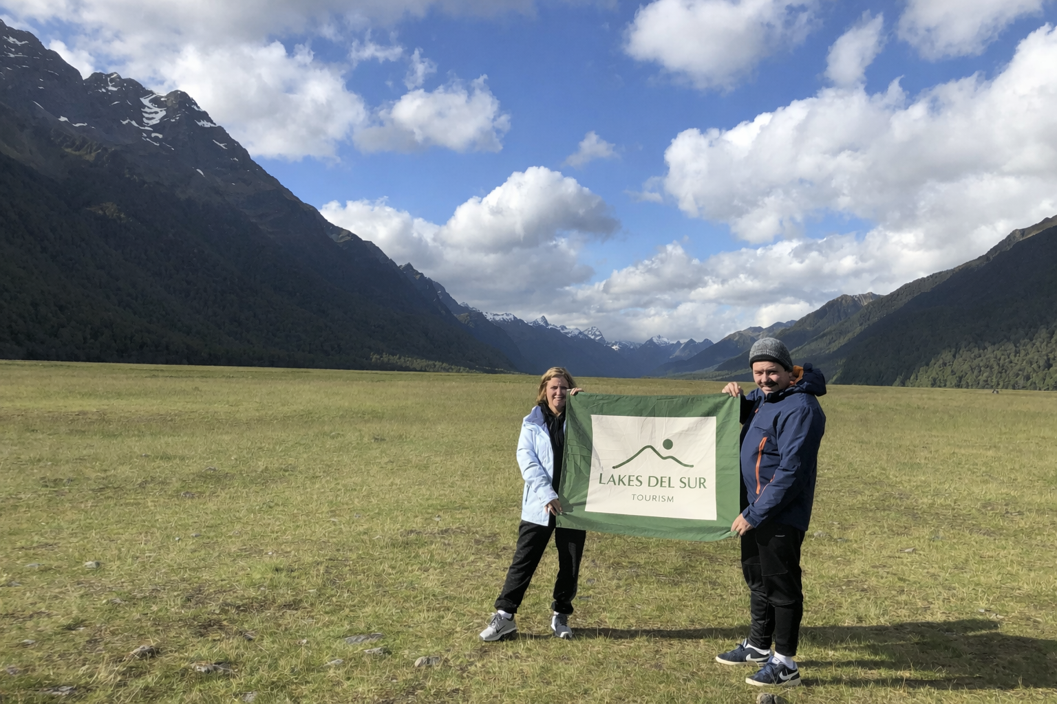 Two people holding a "Lakes Del Sur Tourism" flag in a green field with mountains and cloudy sky in the background.