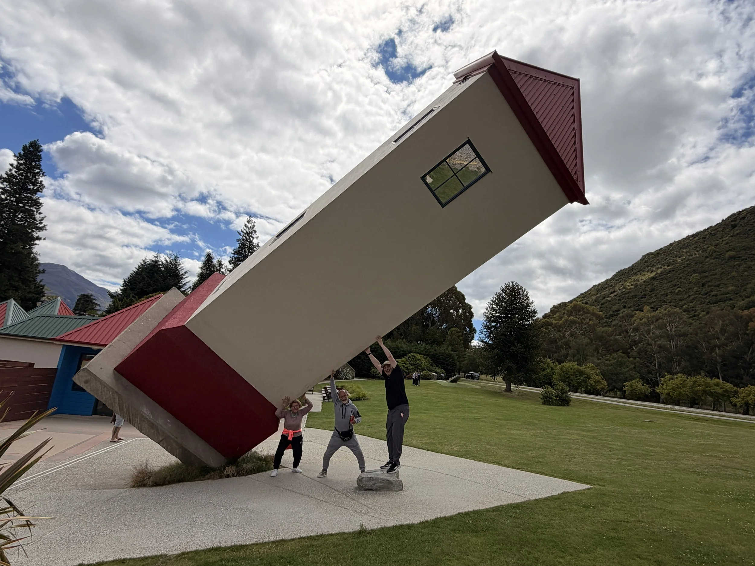 Three people are standing beneath a large upside-down house sculpture outdoors in a park, with mountains and trees in the background, under a cloudy sky.