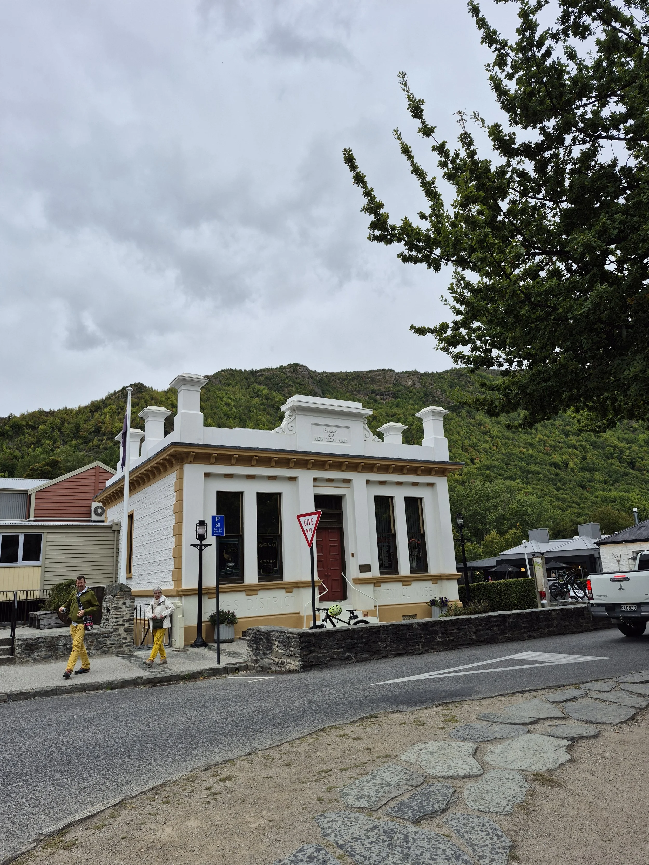 A historic white building with a red door, situated by a street with pedestrians, parked vehicles, and mountains in the background. There are trees and cloudy skies overhead.