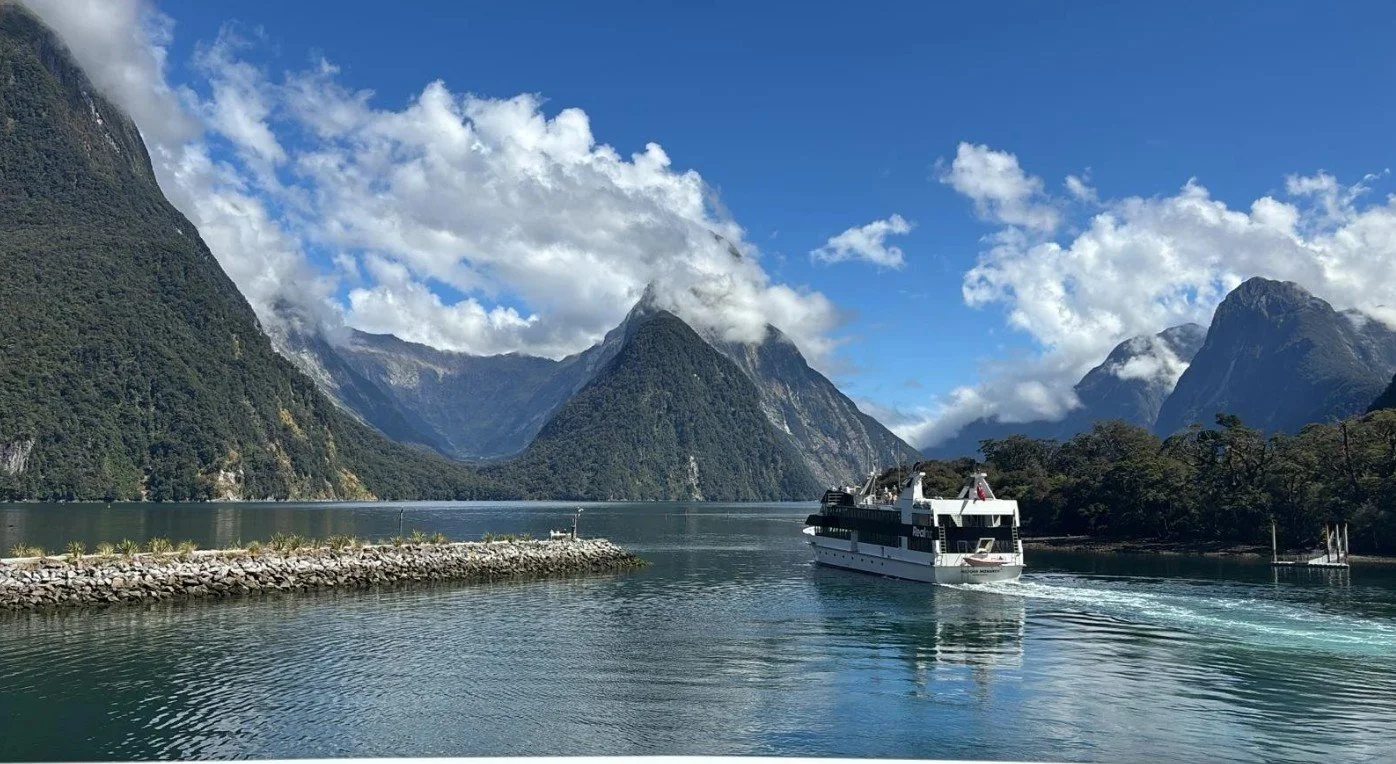 A boat sailing on a fjord surrounded by tall, green mountains with partly cloudy sky.