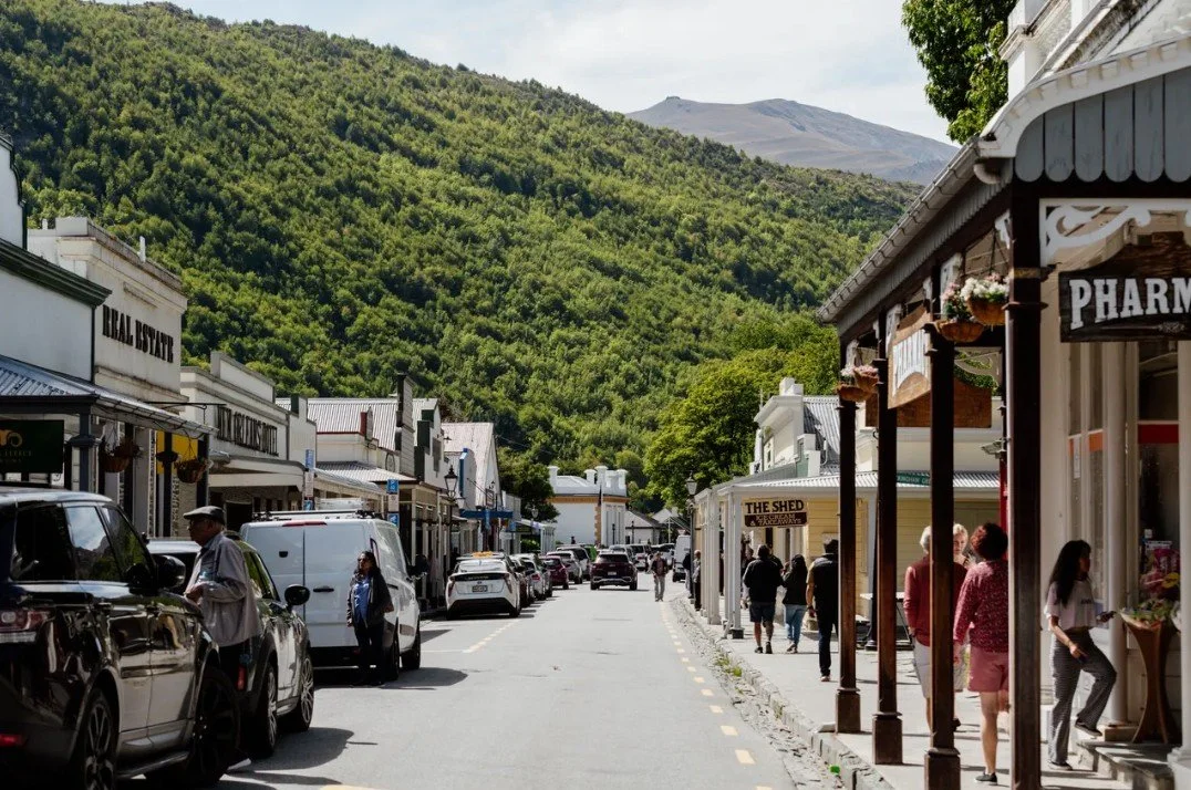 Streetview of a small town with shops and people walking, surrounded by green hillside and mountains in the background.