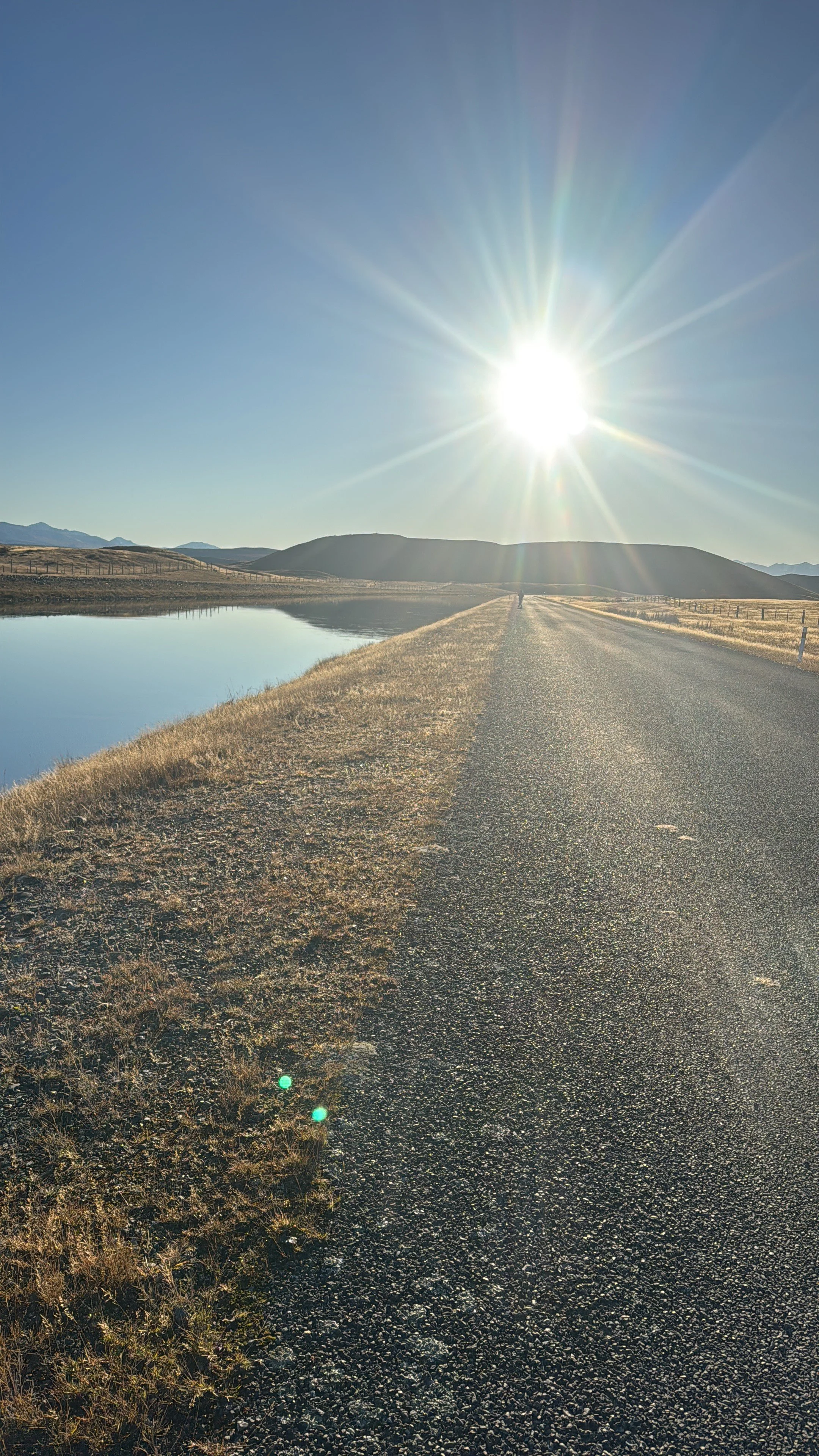 A rural road running alongside a body of water under a bright sun in the sky. Hills are visible in the distance on a clear day.